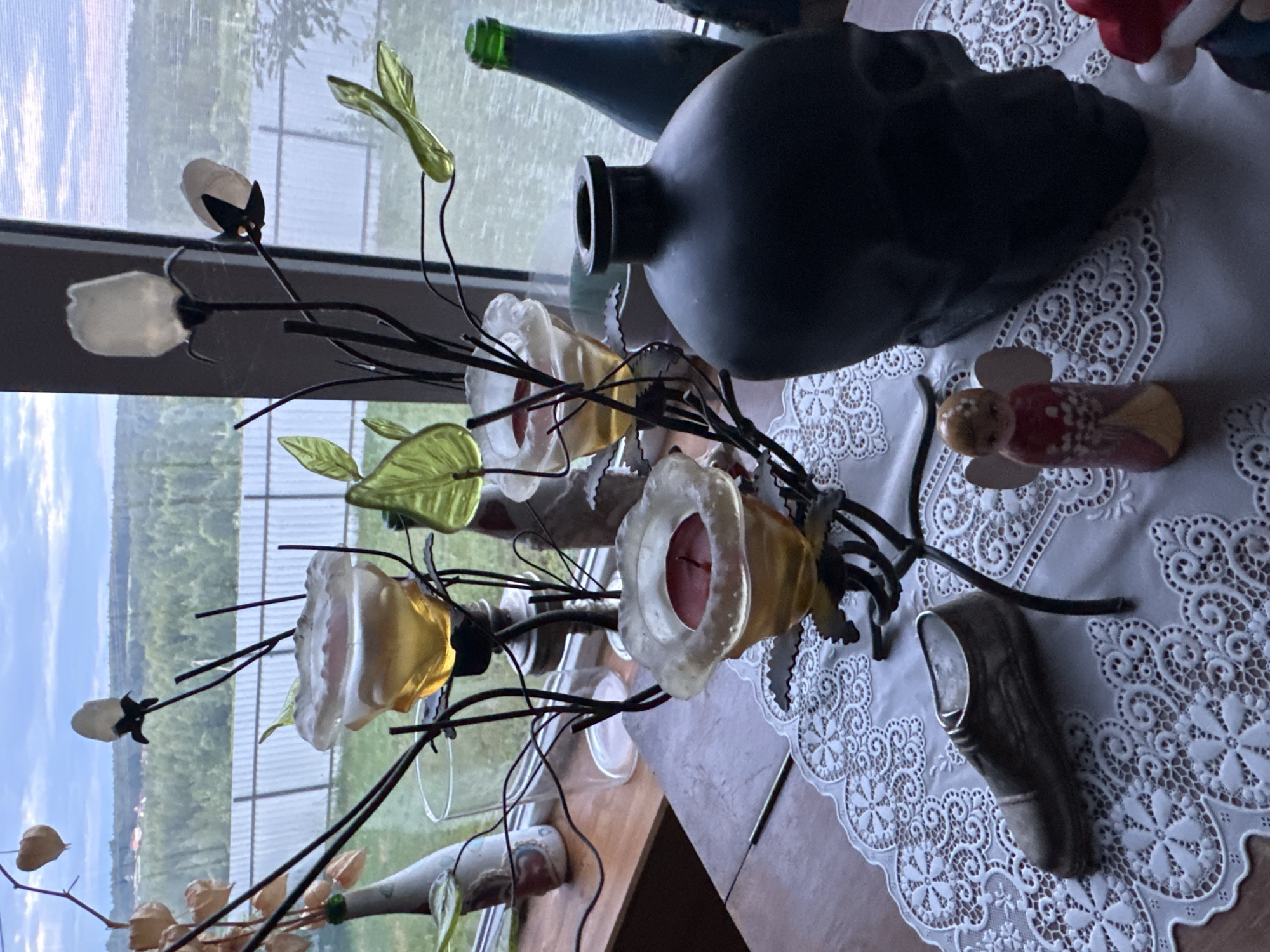 A still life composition is arranged on a wooden table, partially covered by a white lace doily. The arrangement includes a decorative metal candelabra holding three glass candle holders, one with a red candle. To the left of the candelabra is a small, metallic shoe figurine. To the right of the candelabra, a dark, skull-shaped vase dominates the foreground. Between the shoe and the skull vase sits a small, ceramic angel figurine. In the background, through a window, a rural landscape with trees and a fence is visible under a cloudy sky. The scene is indoors, with the window providing a view of the exterior. There are no people or animals present in the image.