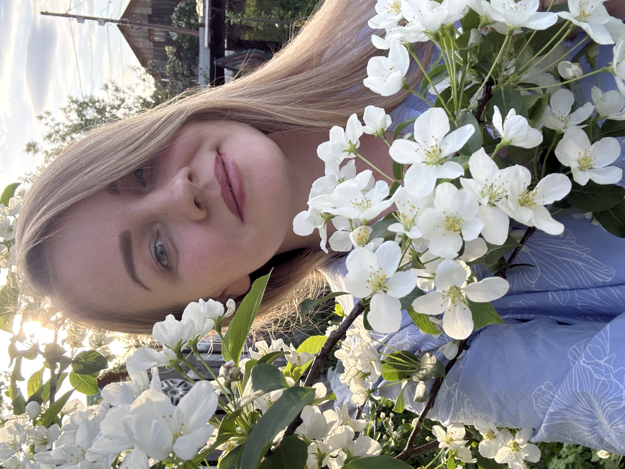 A young woman with light blonde hair is partially visible, her face angled to the left, surrounded by blooming white flowers on a tree branch. She appears to be wearing a light blue shirt with a floral pattern. The background includes a glimpse of a wooden structure, possibly a house, and foliage under a partly cloudy sky. Based on the prompt mentioning the city, it is reasonable to assume this scene is somewhere in Chaikovsky, Russia, possibly a park or garden setting.