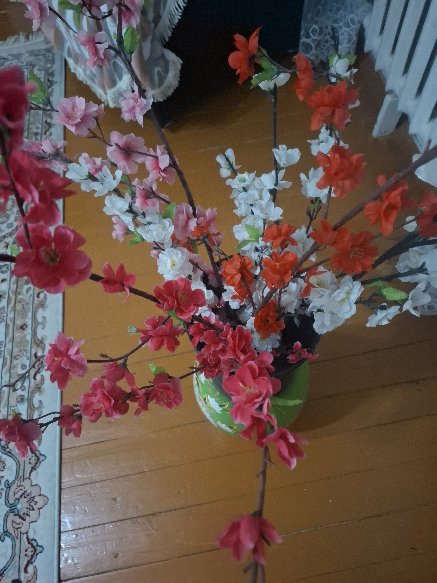 The image shows a close-up of artificial flowers in a vase, arranged on a wooden floor. The flowers are a mix of pink, white, and red, with some green leaves. The vase is green and appears to be ceramic. To the left of the vase, a patterned rug is visible, with a fringed edge. In the background, a white radiator and a draped fabric can be seen. The scene appears to be indoors, in what could be a residential or commercial setting. There are no people or animals present in the image. The lighting suggests it is daytime, possibly with natural light from a window. The style of the rug and radiator might suggest a slightly older building or a particular decor style. The image does not provide any specific clues about the location within the city of Almaty, Kazakhstan, beyond it being an interior scene.
