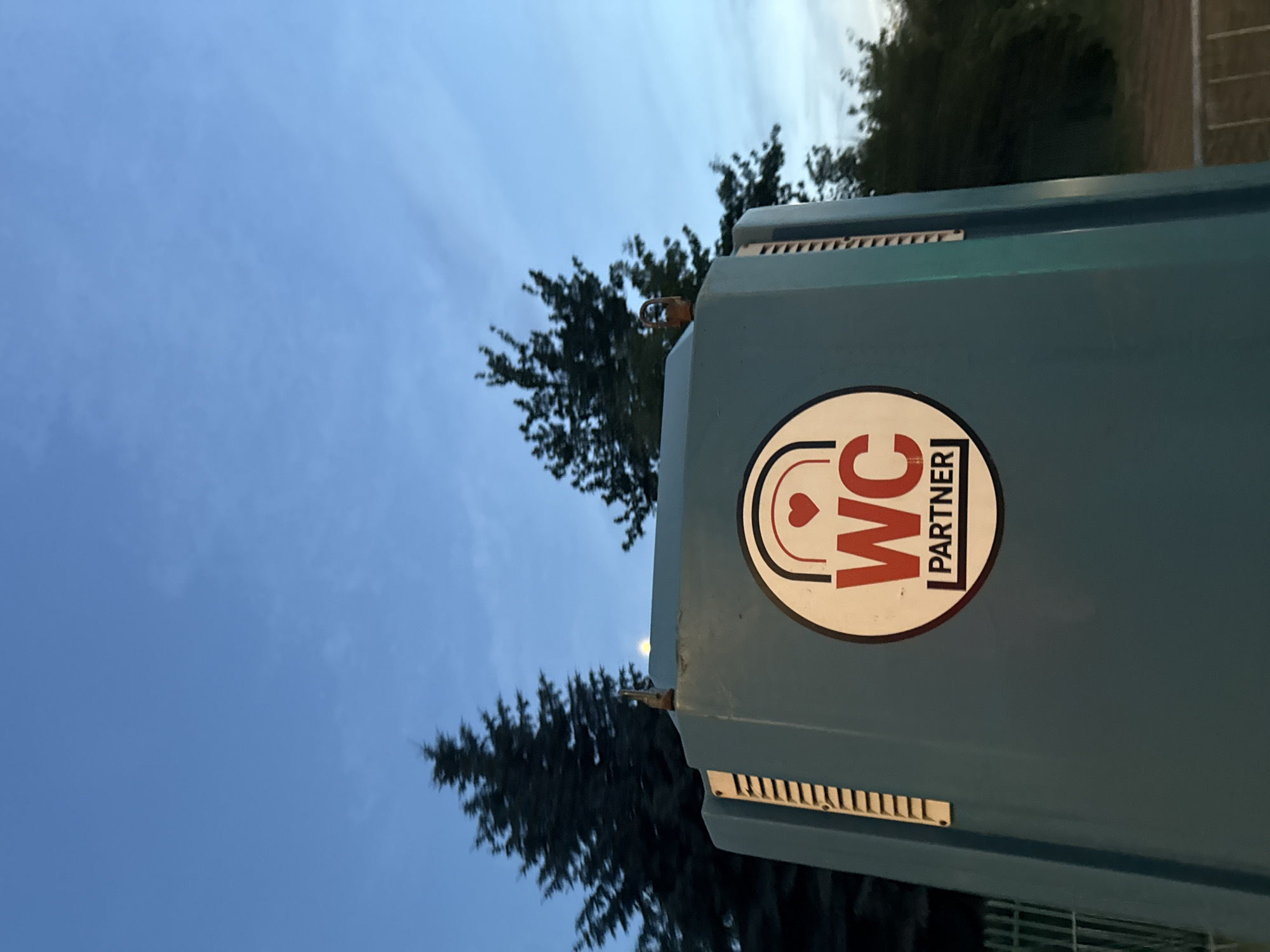 A portable toilet unit with the "WC Partner" logo is visible against a twilight sky with trees in the background. The toilet unit is teal-colored and features a circular emblem with the letters "WC" in red, a red heart above the letters, and the word "PARTNER" in black below. The emblem is framed by black and red curved lines. The toilet unit has vertical vents on its side. Two rusty metal loops are attached to the top of the unit. The sky is a gradient of blue, transitioning from a lighter shade near the horizon to a darker shade above. Tall evergreen trees are visible behind the toilet unit, and a metal fence runs along the bottom of the image.