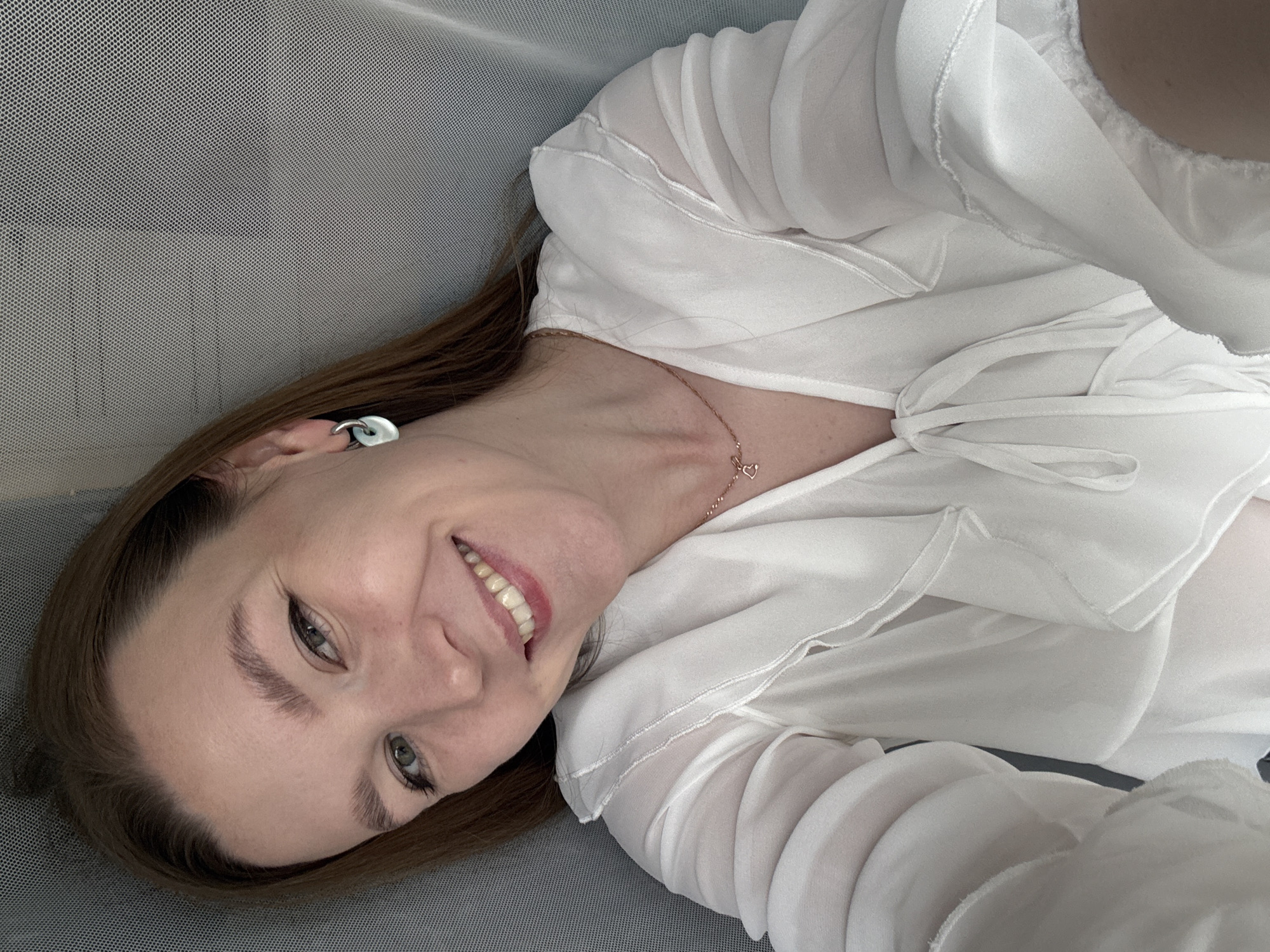 The media file shows a selfie of a woman, appearing to be in her late twenties or early thirties, lying down on a grey, textured surface, possibly a chair or couch. The image is rotated 90 degrees counter-clockwise, meaning the woman's head is to the left of the frame. She has brown hair that falls around her shoulders and is wearing a white, sheer blouse with ruffled details and a tie at the neckline. A delicate rose gold necklace with a heart pendant is visible on her neck. She has light green eyes, accentuated by eyeliner and mascara, and her eyebrows are neatly shaped. Her smile is wide, revealing white teeth, and her lips are painted with a pink lipstick. She is wearing a circular, silver earring in her visible ear. The lighting suggests it is daytime, with soft, diffused light illuminating her face. The background is mostly the grey textured surface, with the sheer fabric of her blouse also creating a significant portion of the frame. There is no visible text in the image. The overall mood conveyed is one of happiness and relaxation. The location context of Izhevsk, Russia, is provided but not visually discernible from the image itself.