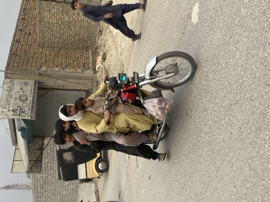 The scene depicts a busy street in Quetta City Tehsil, Pakistan, under an overcast sky. A red motorcycle, license plate "NBT 4902," is moving from left to right, carrying five individuals. An adult male, wearing a light yellow shalwar kameez and a white head covering, drives the motorcycle with a young boy, also in a light brown shalwar kameez, seated in front of him. Behind the driver, a second adult male in a dark brown shalwar kameez and dark head covering is seated, followed by a young male in a similar dark brown shalwar kameez. A fifth young male, wearing black shalwar kameez, stands on the rear foot pegs. A clear plastic bag containing red onions hangs from the front of the motorcycle.
In the background, on the left, a light-colored auto-rickshaw is partially visible near a grey concrete block wall. Adjacent to the wall is a building with a closed metal shutter and a blue awning. Above the shutter, a white sign with Urdu script and contact numbers ("0313-8983338," "0315-8853518") identifies a "Mobile & Motorcycle Repair Shop." A cellular tower is visible in the distant background.
To the right, a young male in a dark blue shalwar kameez and sandals walks leftward, carrying a light shoulder bag, observing the motorcycle. Behind him, a brick-walled building with a closed gate is visible. A light-colored dog lies on the dusty ground next to the building. The road surface is a speckled grey asphalt or concrete.