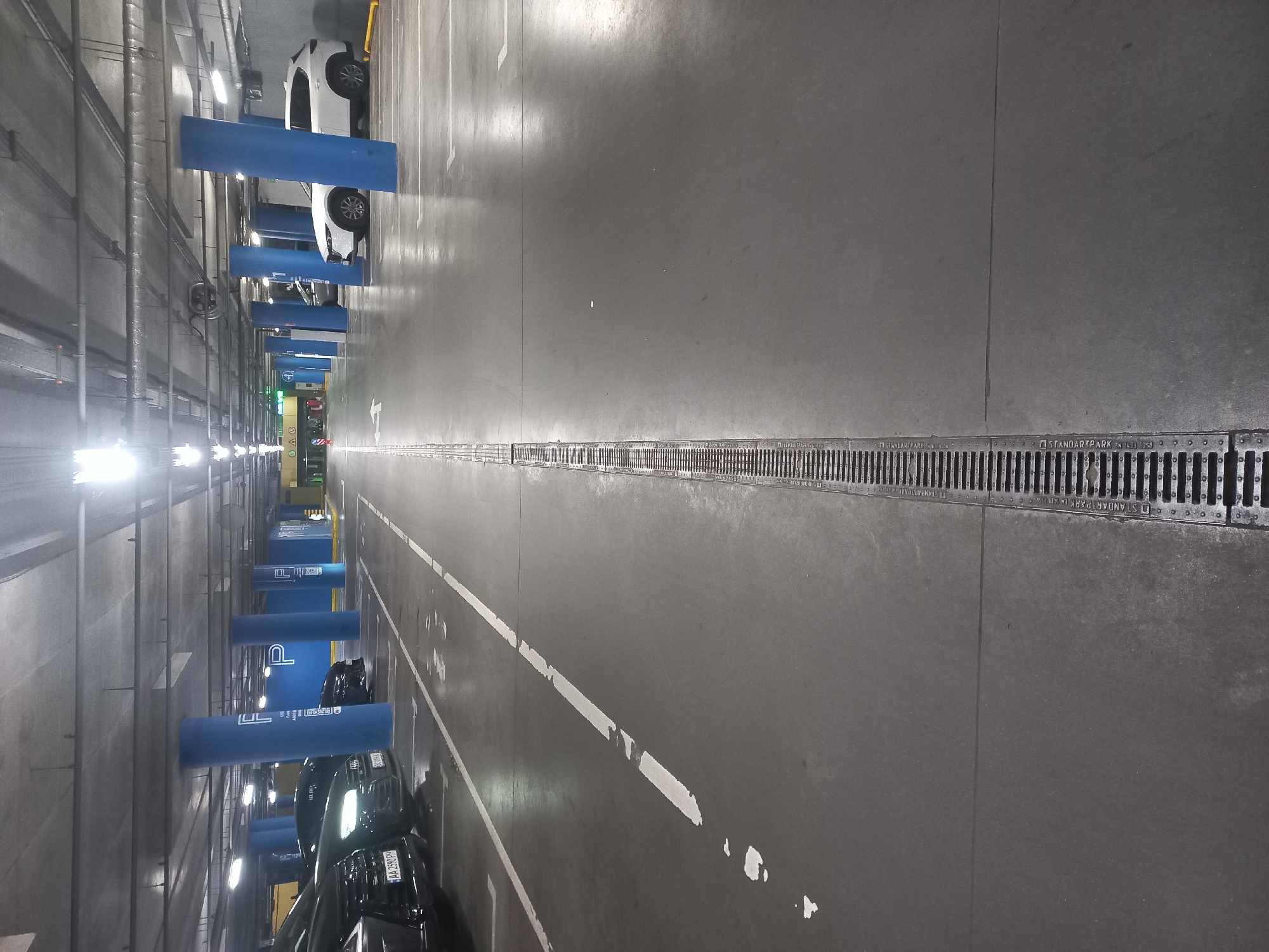 This media file is a vertically oriented photograph of an underground parking garage in Kyiv, Ukraine. The image captures a long, brightly lit aisle with parked cars and infrastructure visible.
On the left side of the frame, several vehicles are parked. A white SUV is visible near the top, and further down, multiple darker-colored cars are parked. The visible Ukrainian license plates, such as "AA 2530HP," "AA 2380HP," and "AA 2590HP," confirm the Kyiv region ("AA" series). These cars are parked next to a series of blue, cylindrical barriers or pillars, some of which bear the partial text "UL."
The floor is concrete, marked with white lines delineating parking spaces and directional arrows in the driving aisle. A prominent metal grate drain, spanning the length of the visible aisle, runs down the center of the driving lane. Text "STANDARTPARK EN 1433" is discernible on these drain grates in several places.
Overhead, artificial fluorescent lights illuminate the space, and exposed pipes, ventilation ducts, and electrical conduits are visible on the ceiling. In the far distance, various blurry signs and lights are visible, including what appears to be a green exit sign. The scene depicts a typical, orderly underground parking environment, with no people visible and no specific event occurring beyond the cars being parked. The lighting suggests an indoor setting, making the time of day irrelevant.