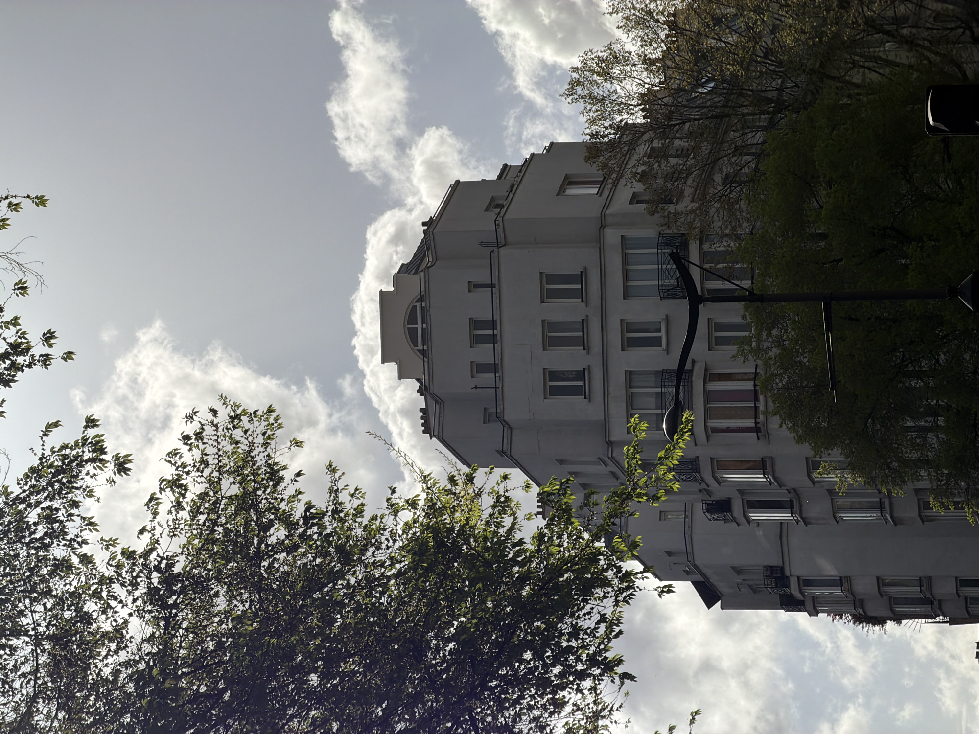 The image shows a tall, white building in Paris, France, partially obscured by trees. The building has multiple windows and balconies, with an unusual stepped roofline. The sky is overcast with some visible clouds. A streetlight is also visible on the right side of the image.