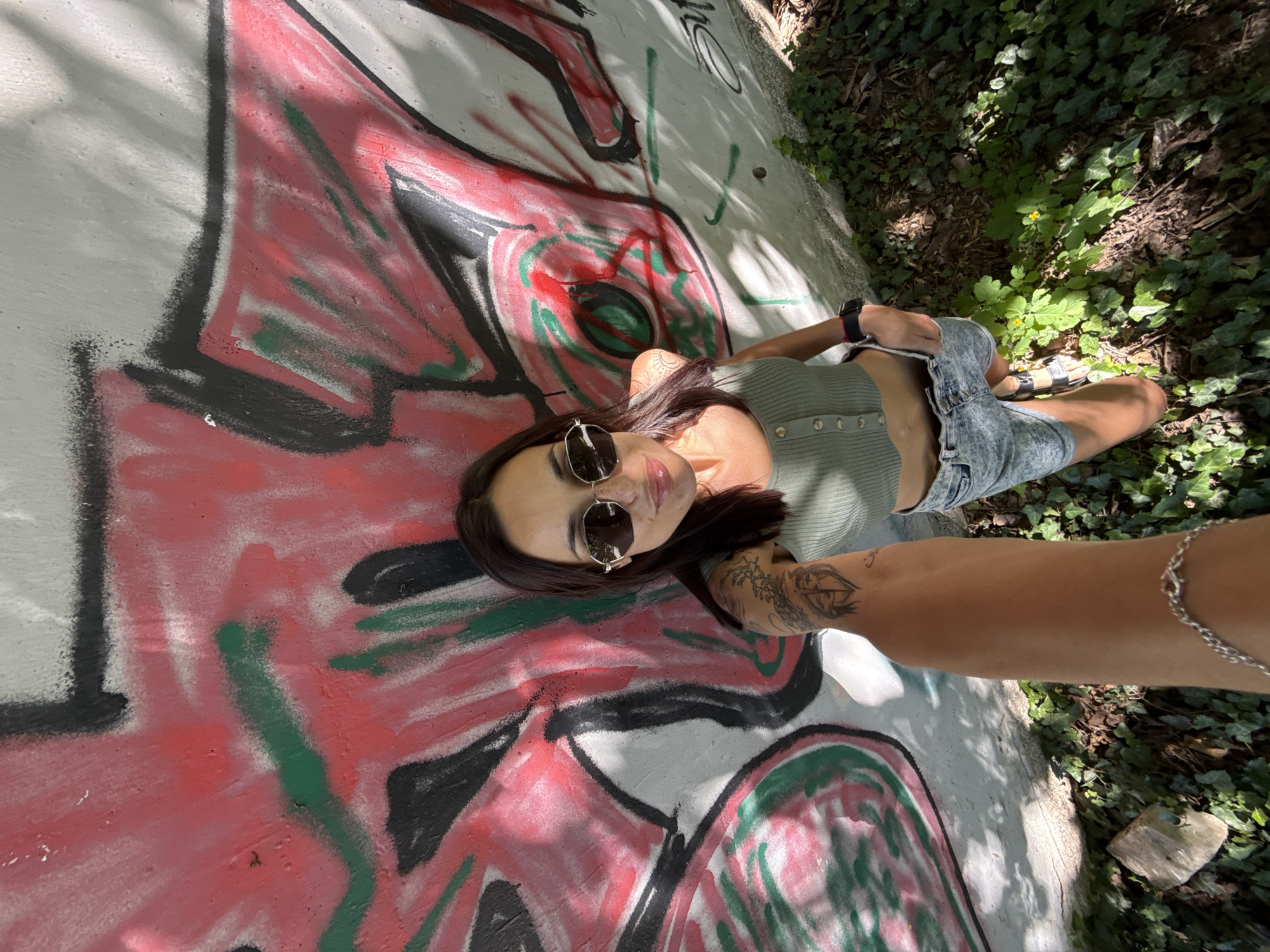 The image shows a woman lying on a concrete surface covered in graffiti, located next to an area of greenery. She is wearing sunglasses, a light green top, and denim shorts. The woman is holding her arm out towards the camera, revealing a tattoo on her forearm and a silver chain bracelet. The graffiti consists of red, black, and green paint on a white background. The greenery includes leaves and small yellow flowers. The location appears to be outdoors, adjacent to a wall or structure. Pleven, Bulgaria cannot be ascertained from the image.