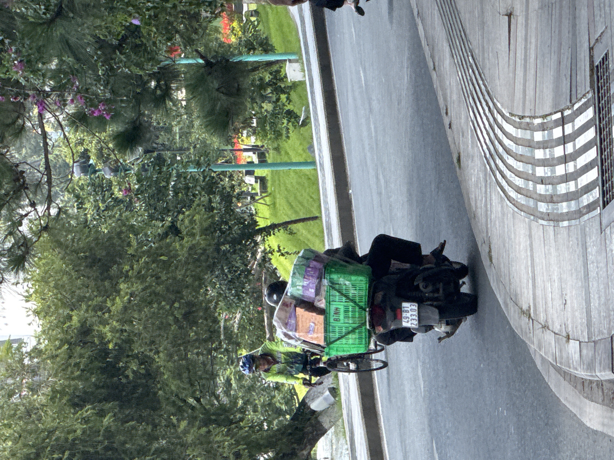 In Phường Xuân Hương - Đà Lạt, Vietnam, a person on a scooter is carrying a large green basket filled with various items, including what appears to be packaged goods and possibly other deliveries. The scooter has a license plate visible that reads "49-B1 233.03". Another person is riding a bicycle in the background, wearing a bright yellow and green jacket and a blue helmet. The scene takes place on a paved road with a curved sidewalk. Lush green trees and foliage are present on either side of the road, with some flowering plants visible in the background. The overall environment suggests an outdoor, urban setting.