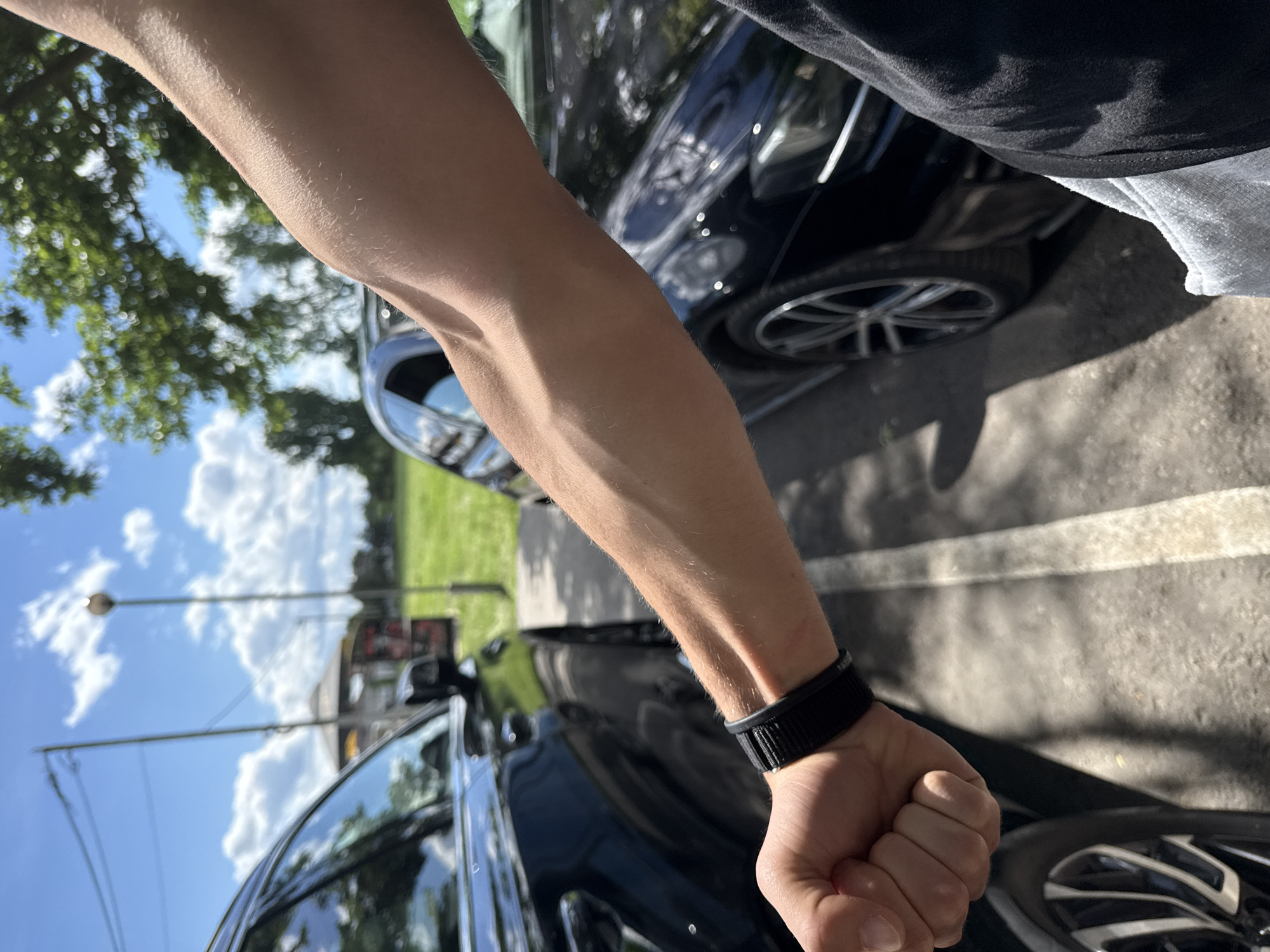 A man's forearm with visible veins is positioned in front of a black car. The man is wearing a black wristband. The background includes a blue sky with clouds, trees, a green field, a building with a sign, and a street lamp. The photo appears to be taken outdoors in a residential area. There is no evidence to suggest that it is in Elhovo, Bulgaria.