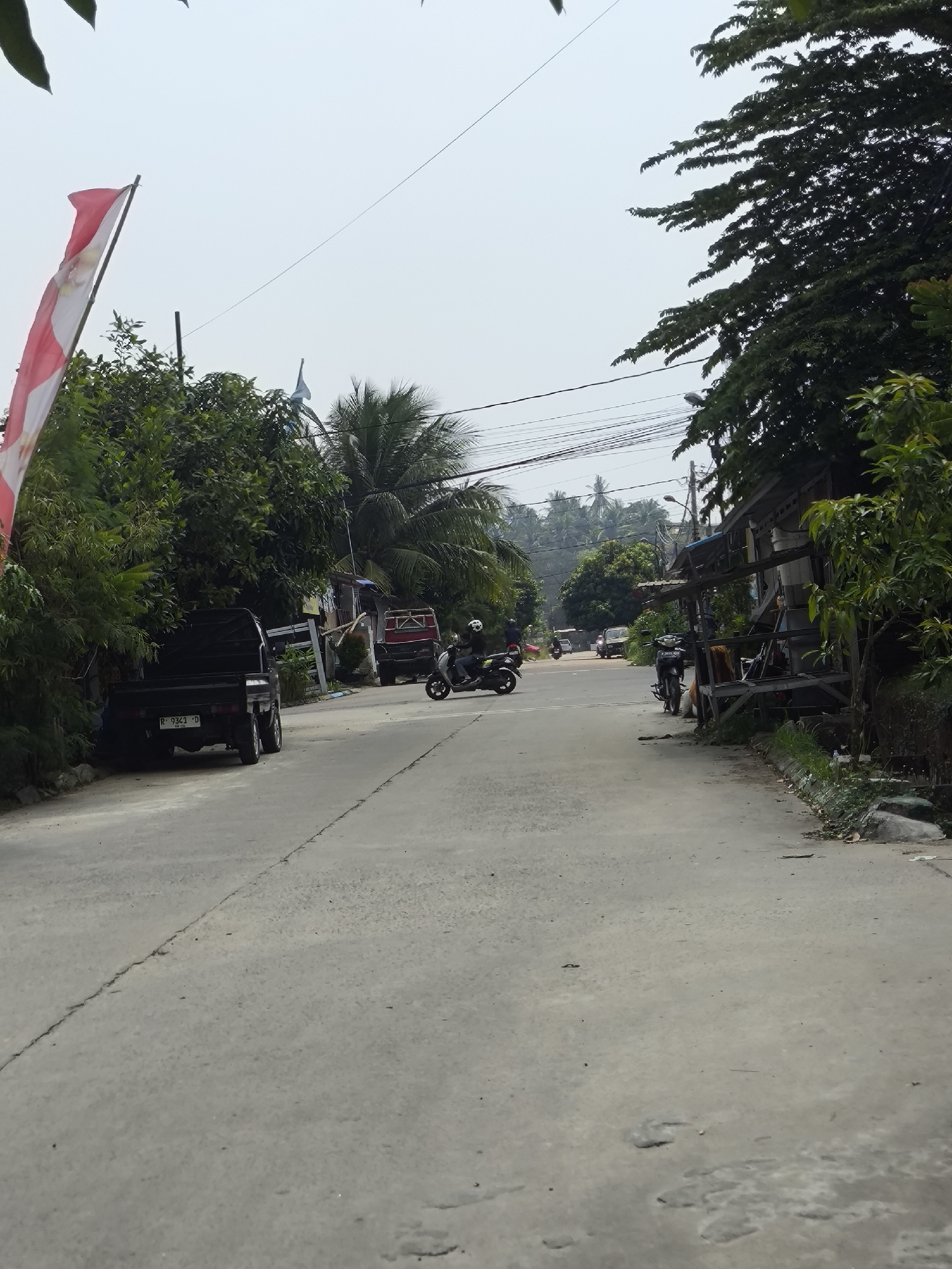 A sunny day in Cilegon, Indonesia, shows a paved road with a faint white line down the middle. On the left, a black pickup truck with license plate R 9343 D is parked. Beside it, lush green trees and foliage line the roadside, with a red and white Indonesian flag fluttering on a pole. Further down the road, a red truck is visible, and a person on a black scooter with a white helmet rides past. On the right side of the road, a simple wooden structure with a thatched roof sits adjacent to more green trees and a few small rocks. In the distance, more palm trees and houses indicate a residential area, with power lines strung across the hazy sky.