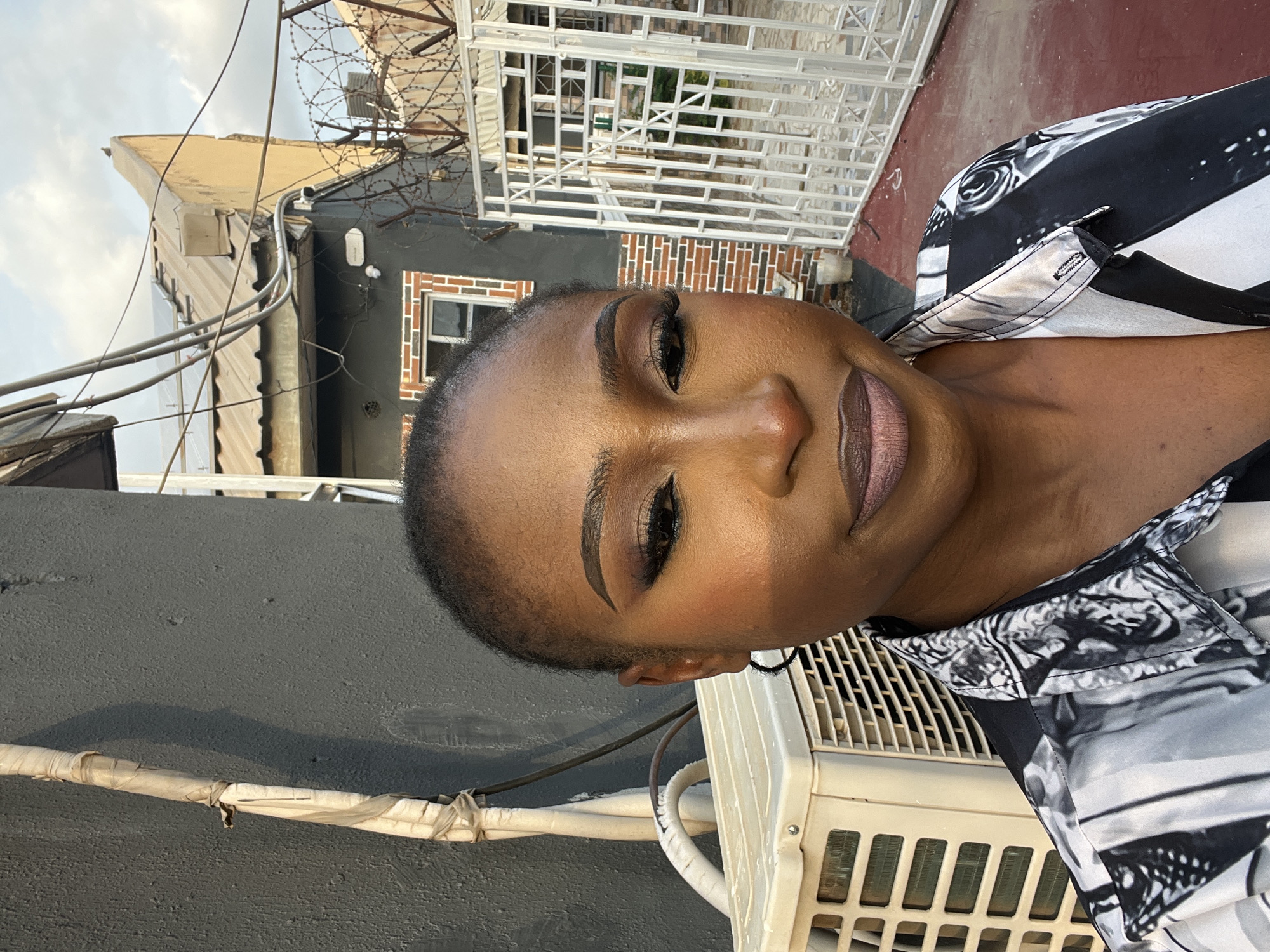 A headshot captures a Black woman with a short buzz cut and full makeup, including bold eyeshadow and lipstick. She is outdoors in what appears to be a courtyard. To her left, a beige outdoor air conditioning unit is visible. Behind her, a grey textured wall is present, with white piping running vertically along it. Further back, a collection of buildings with corrugated metal roofs, visible power lines, and barbed wire fencing suggest an urban setting. A white metal gate with intricate geometric patterns is on the right side of the frame. The ground is a reddish-brown color. The lighting suggests it is late afternoon, casting shadows on the wall.