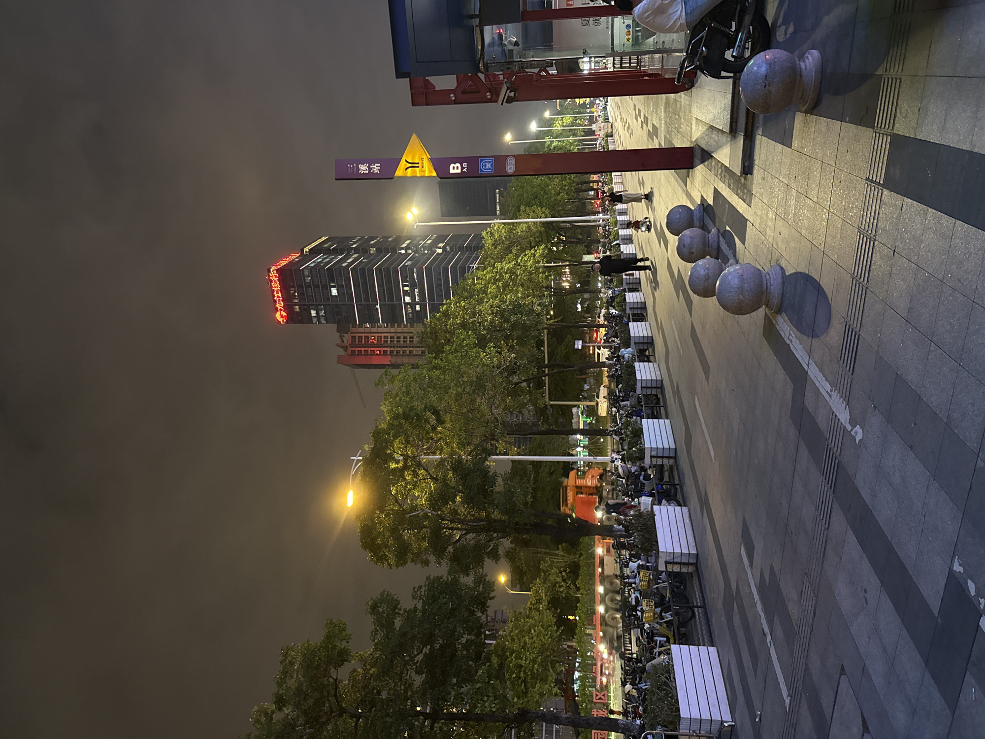 The image captures a nighttime street scene in Tianhe District, China. A tall, modern building with illuminated signage in Chinese characters and a red border is visible in the background. Adjacent to it is another tall building. In the foreground, a street is lined with parked motorcycles and bicycles. Along the sidewalk, there are planters with greenery and large spherical stone bollards. A purple signpost with Chinese characters and a yellow directional arrow indicates "三溪站" (Sān xī zhàn), likely a station name, and the letter "B" with an entrance symbol. Several people are walking on the sidewalk, including a man in a dark shirt and pants, a woman in a light-colored top and dark pants, and a child on a small ride-on toy. Another person is seen from behind sitting on a motorcycle. The street is paved with grey tiles and has white lines. Streetlights illuminate the scene.