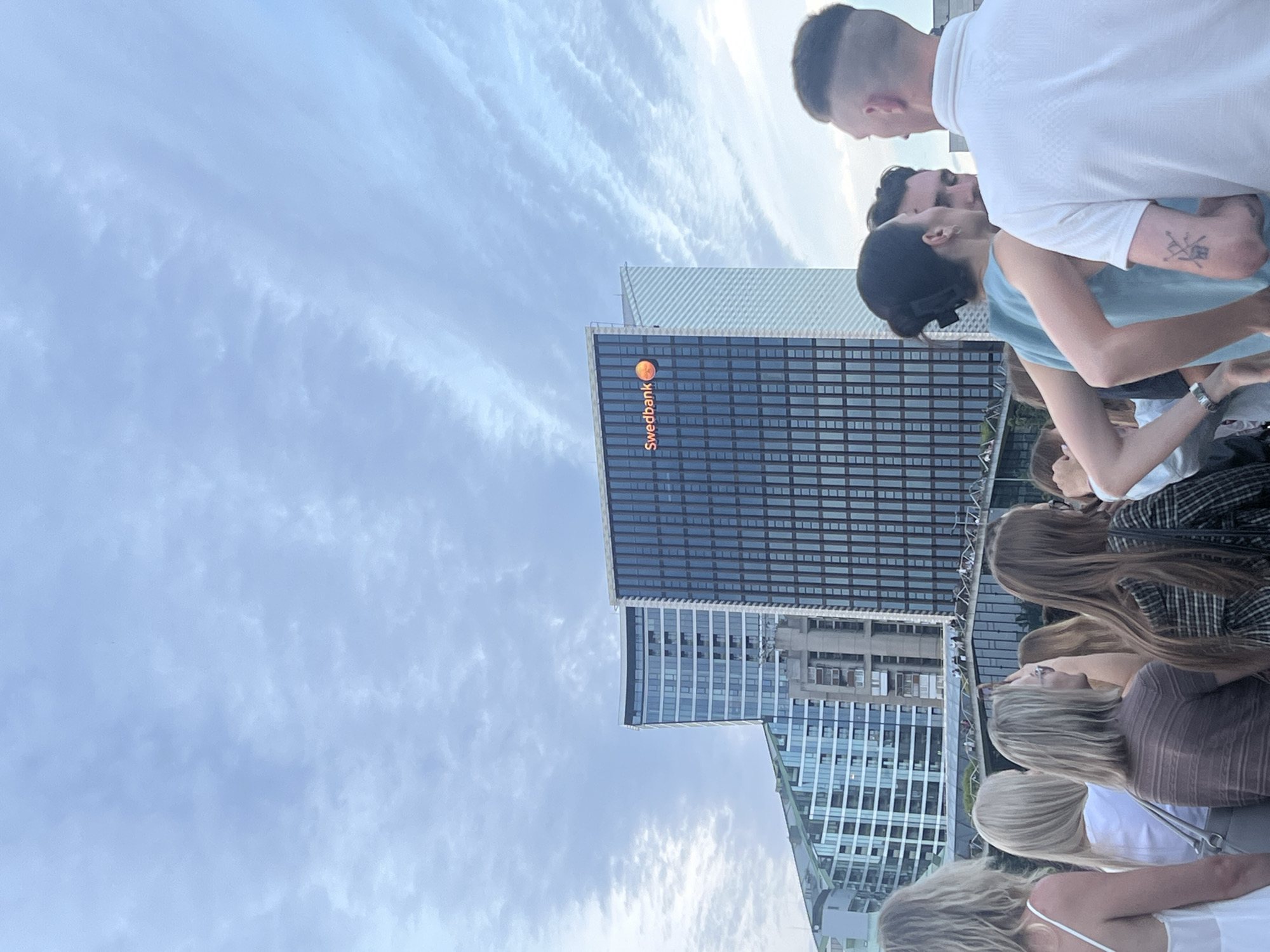 The image captures a crowd of people standing on a pedestrian bridge in Vilnius, Lithuania, with modern skyscrapers in the background. In the center of the frame, a tall, dark glass building prominently displays the "Swedbank" logo and an orange circular emblem. To the left, another contemporary building features a distinctive curved facade with numerous windows. The sky above is filled with scattered, wispy clouds. The individuals in the foreground are facing away from the camera, engaged in conversation and looking towards the cityscape. One person, a man on the right, is wearing a white polo shirt and has a distinctive haircut. A woman in the center is wearing a teal sleeveless top and has her hair pulled back in a bun. The overall scene suggests a gathering or event taking place in a developed urban environment.
