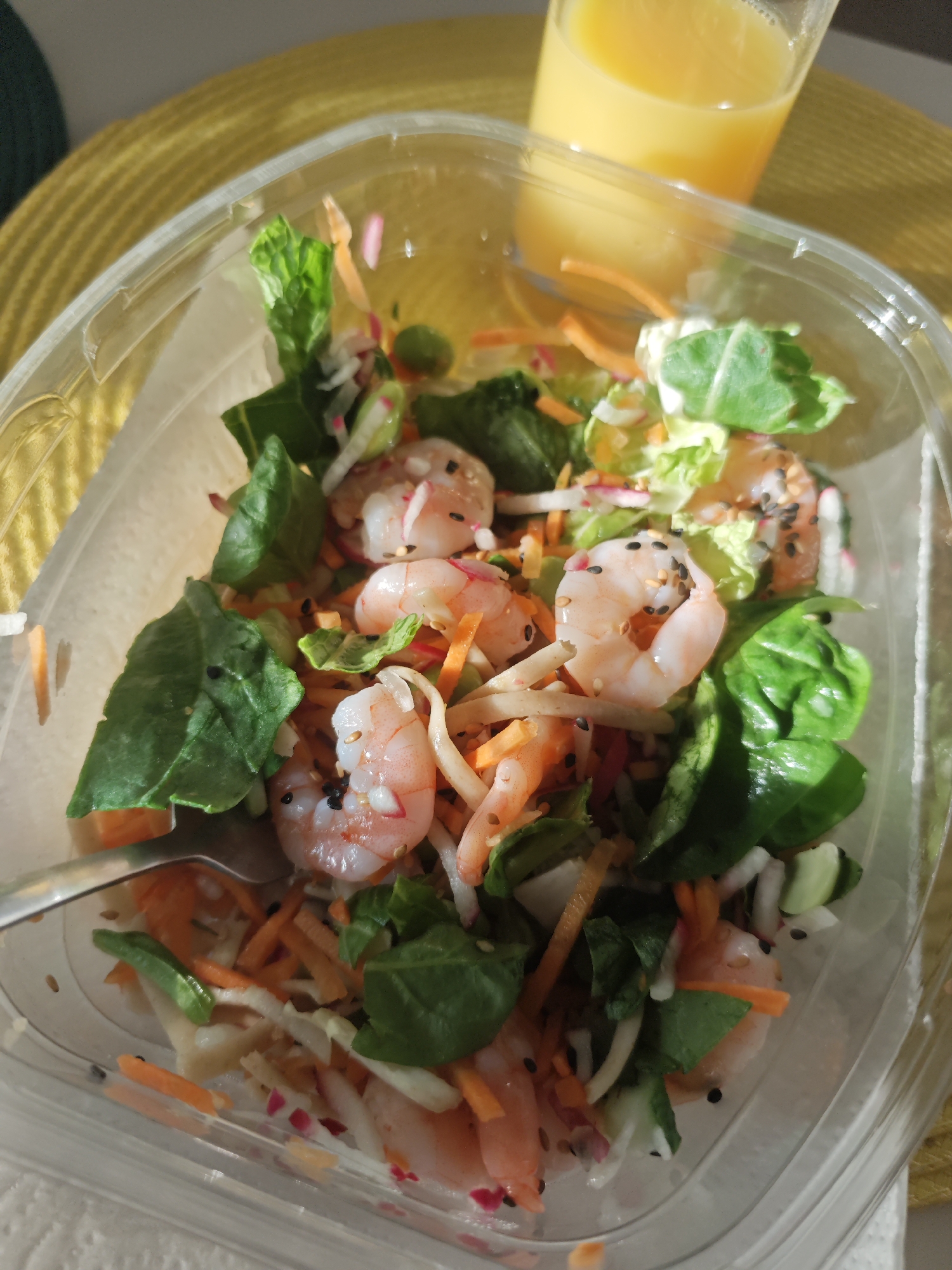 The image shows a close-up, overhead view of a shrimp salad in a clear plastic takeout container, placed on a yellow placemat. A glass of orange juice is visible in the upper right corner of the frame. The salad contains cooked shrimp, shredded carrots, leafy greens, and possibly radish and other finely chopped vegetables, garnished with black sesame seeds. A fork is partially visible on the left side of the container, resting on the salad. The lighting suggests it is daytime, possibly a meal being consumed outdoors or near a window, with sunlight casting shadows. There are no people or animals in the image. The setting appears to be a casual dining environment. The image was not taken in Tilburg, Netherlands.