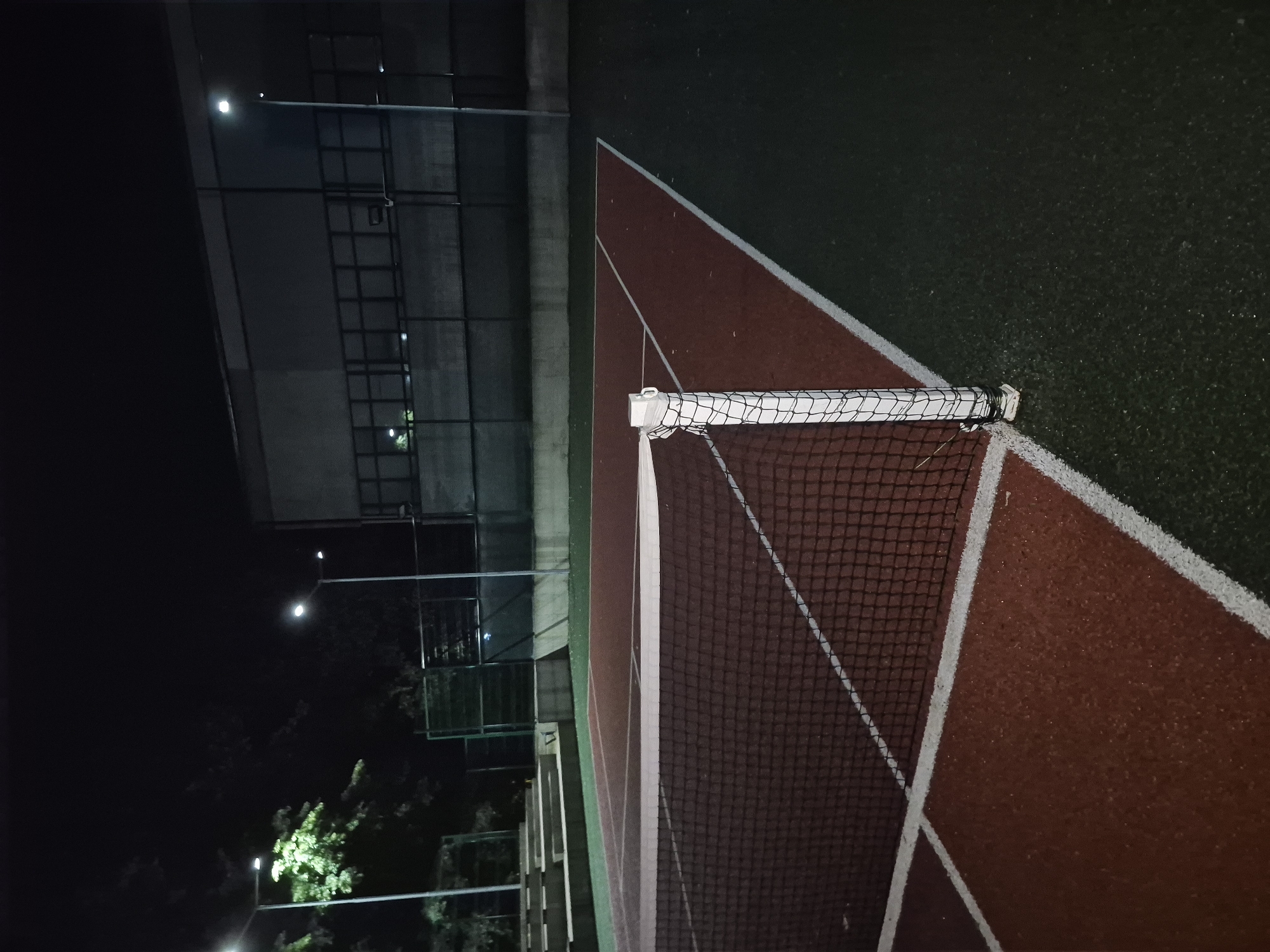 This is a nighttime photograph of an outdoor tennis court in the city of Popovo, Bulgaria. The court has a red playing surface with white boundaries and is surrounded by green artificial turf. A tennis net with a white top band is visible in the foreground, supported by a white pole. In the background, there are tiered concrete bleachers, a building with a dark facade, and several lampposts illuminating the area. Trees with green foliage are also visible to the left. The scene is deserted, with no people present.