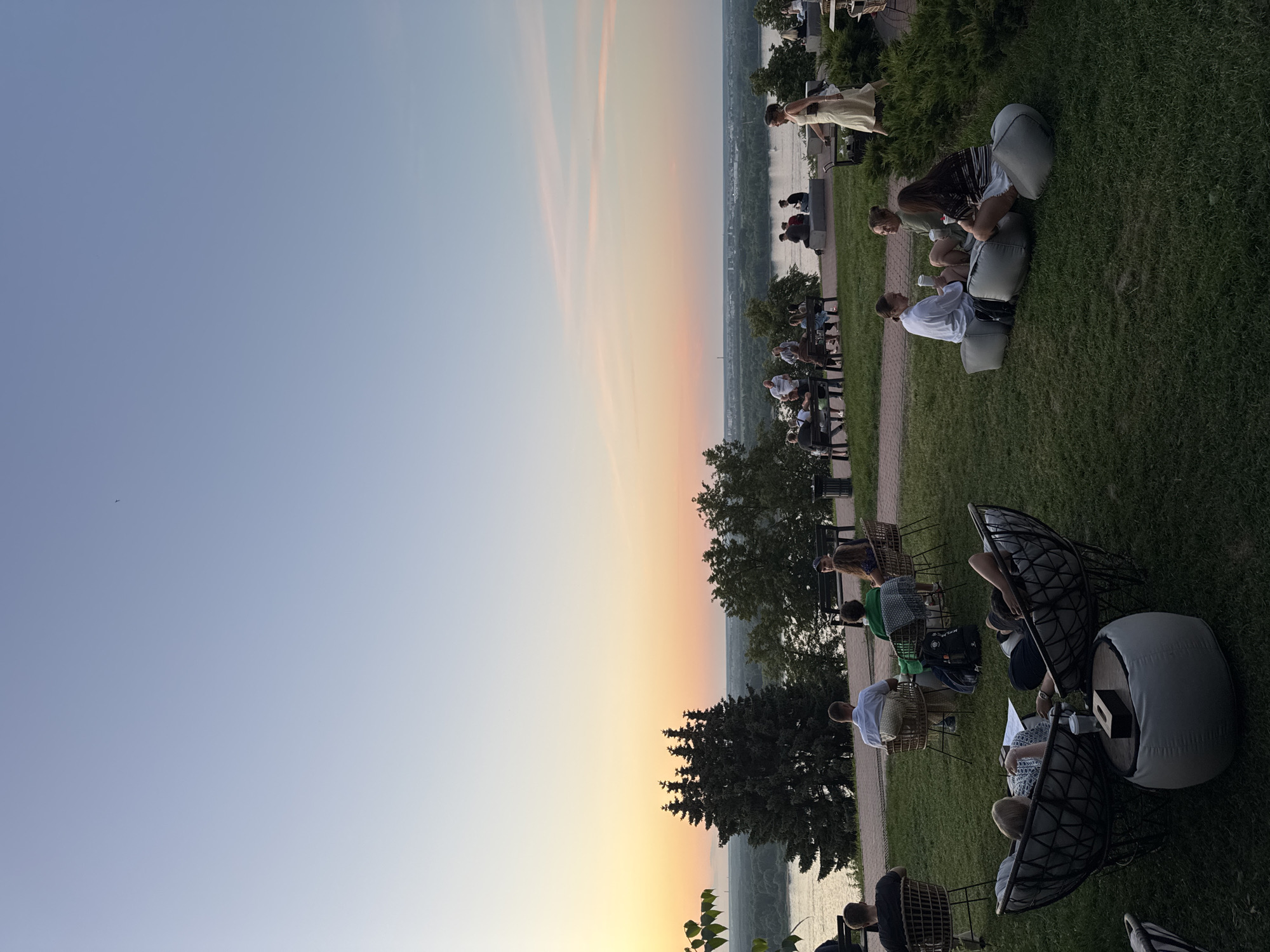 The image depicts a panoramic view of the city of Nizhny Novgorod, Russia, during sunset. The foreground is occupied by a green lawn with several people relaxing on beanbag chairs and wicker chairs. A low table with a tissue box sits between two large wicker chairs in the center-left foreground. In the middle ground, a group of people are seated on benches and chairs, overlooking a river. The river, with a cityscape in the distance, is visible behind a line of trees. The sky displays a gradient of colors, transitioning from a light blue at the top to soft orange and pink hues near the horizon, indicating the sunset. A lone woman in a light-colored dress walks across the scene in the middle ground. The overall atmosphere suggests a serene evening gathering at a scenic overlook.
