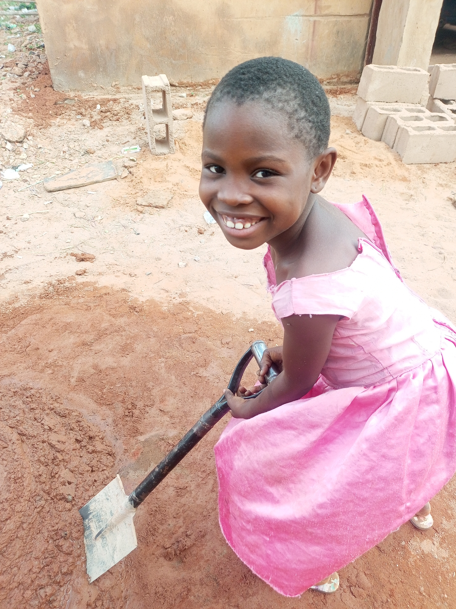 A young African girl, approximately 5-8 years old, is depicted in a medium shot, beaming brightly at the camera. She has short, dark hair and is dressed in a vibrant, off-the-shoulder pink dress, which appears to be made of a satiny fabric with ruffled or pleated details on the back. She is crouching or kneeling on reddish-brown earth, holding a metal shovel with a black handle, the blade of which is partially submerged in a patch of wet, muddy ground directly in front of her.
The scene is an outdoor, unpaved area with distinctive reddish-brown soil, characteristic of regions like Tempure, Nigeria, the provided location context. In the background, a large, light brown, cylindrical structure, possibly a water tank or cistern, dominates the left side. To the right, several light grey concrete blocks are visible, some stacked and others lying on the ground, suggesting a construction or utility area. Minor debris, including small rocks, is scattered across the ground.
The girl's posture and joyful, wide smile suggest she is playfully engaging with the shovel and mud. The photograph was taken during daylight hours under clear weather conditions, indicated by the bright and even lighting. There is no visible text in the image.