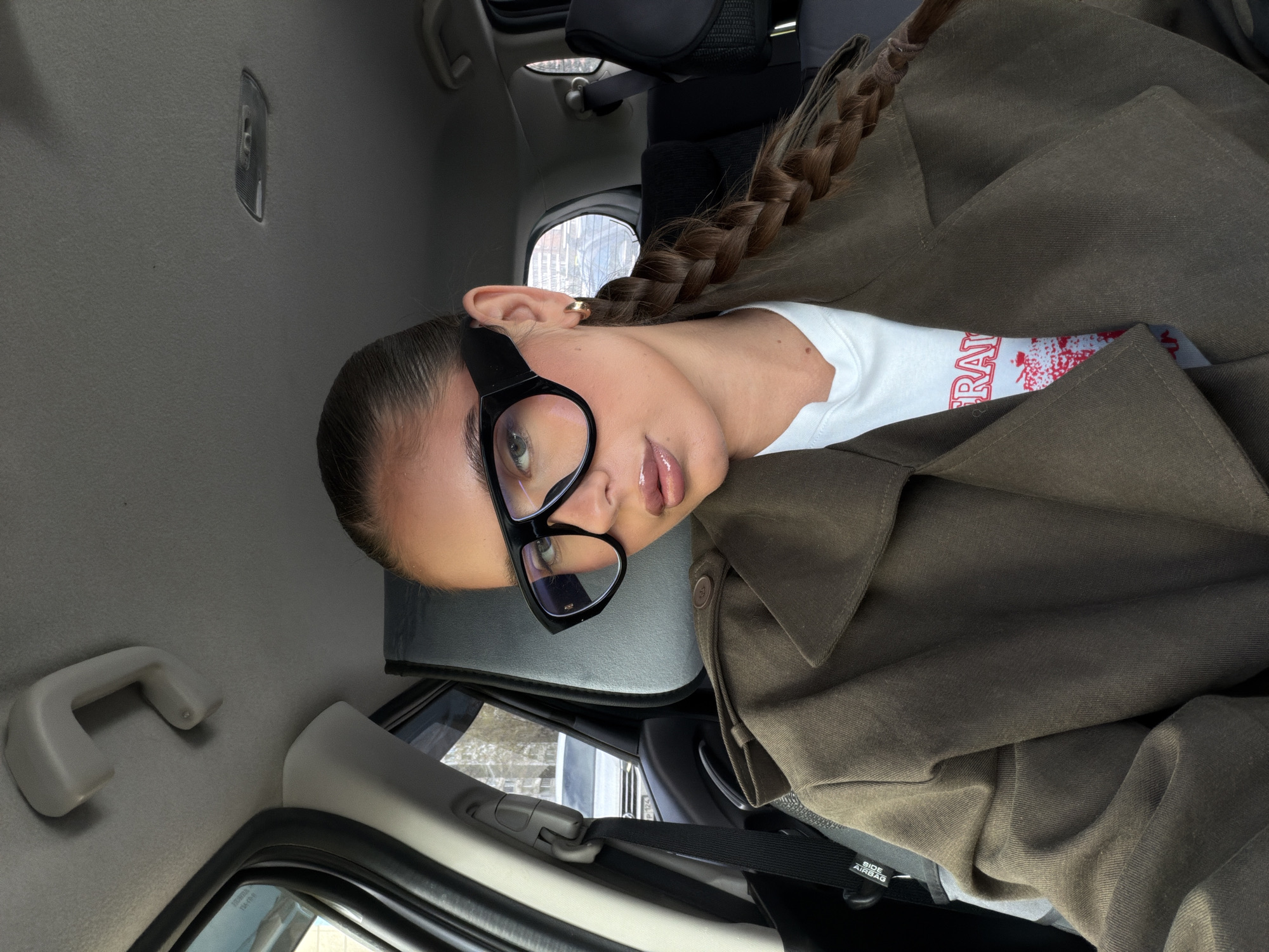 A young woman sits in the passenger seat of a car. She wears large black glasses and a white t-shirt with red lettering, layered under an olive green jacket. Her hair is styled in a single braid. The car's interior is visible, including the roof, window, and door handle. The vehicle appears to be in the city of Krasnoyarsk, Russia, as evidenced by the building visible through the window.