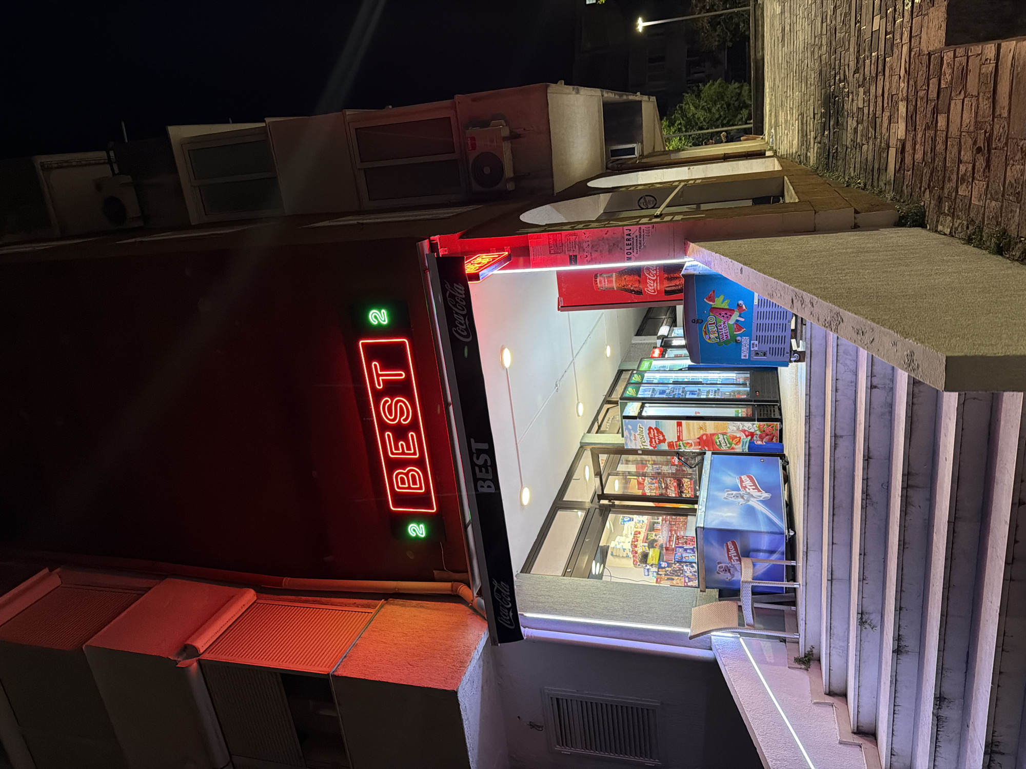 A night exterior shot depicts a convenience store named "BEST" with neon signage. The store is located at street level, with a set of concrete stairs leading up to its entrance. A light blue cooler with a "Frikom" logo is situated on the steps. Inside the store, shelves are stocked with various products. A Coca-Cola advertisement is visible on a red vertical display near the entrance. The building has multiple floors with windows and balconies. A street lamp illuminates the cobblestone sidewalk to the right of the store.