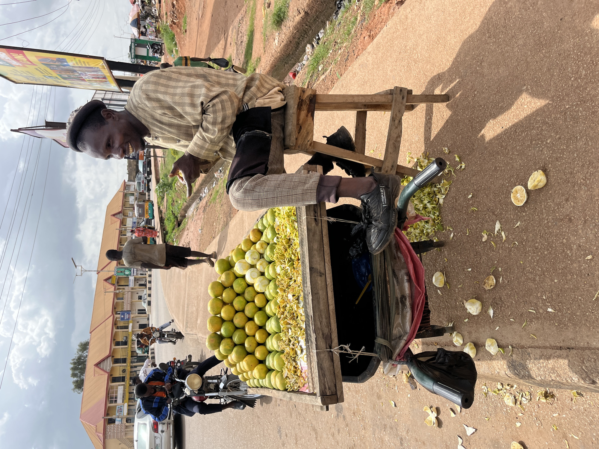 A man is sitting on a wooden stool next to a cart filled with oranges. He is wearing a checkered smock and pants and a kufi hat. He is holding an orange in his left hand. The cart is made of wood and is attached to a bicycle. The scene appears to be a street market in Gura Top, Nigeria. The street is paved with asphalt, and there are buildings in the background. The sky is cloudy. The man is smiling.