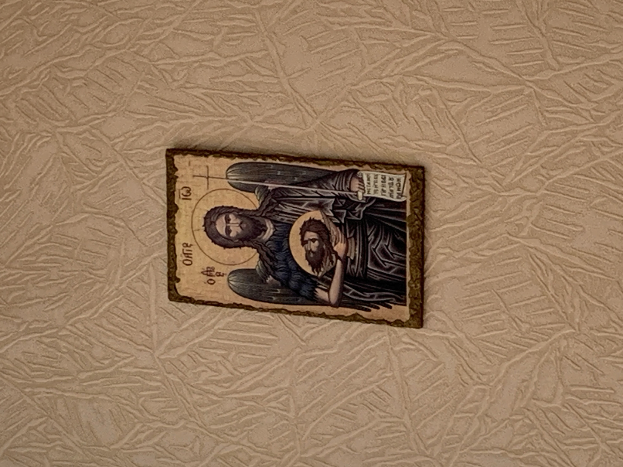 A close-up shot depicts a small, rectangular, framed icon resting on a textured, beige surface. The icon portrays a winged figure, likely an angel or saint, holding a severed head. Cyrillic script is visible on the icon. The textured surface on which the icon rests appears to be wallpaper or a similar wall covering. There is no other information provided regarding the city of Chaikovsky, Russia.