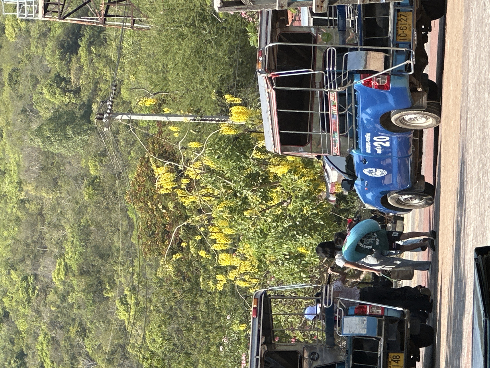 A daytime shot shows two people in motion on a Pattaya City, Thailand street near an open-air taxi. A lush, mountainous forest landscape is behind the street. The people are walking between two parked open-air taxis, with one person carrying an inflated blue inner tube. The taxis are blue and silver with black interior and metal bars. The taxis have license plates. A third vehicle is partially visible on the right edge of the frame. Trees with yellow flowers are abundant. A transmission tower is at the top of the frame.