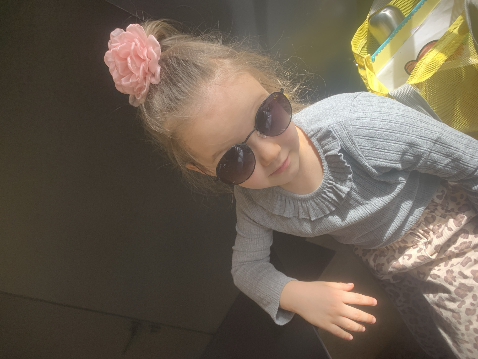 A girl is positioned above the camera, looking down with sunglasses on. She has a pink flower accessory in her hair. The girl wears a gray long-sleeved shirt and patterned pants. Part of a yellow mesh bag is visible in the upper right corner. The lower left of the image is dark, suggesting an indoor setting. There is no information available to determine the location in Izhevsk, Russia.