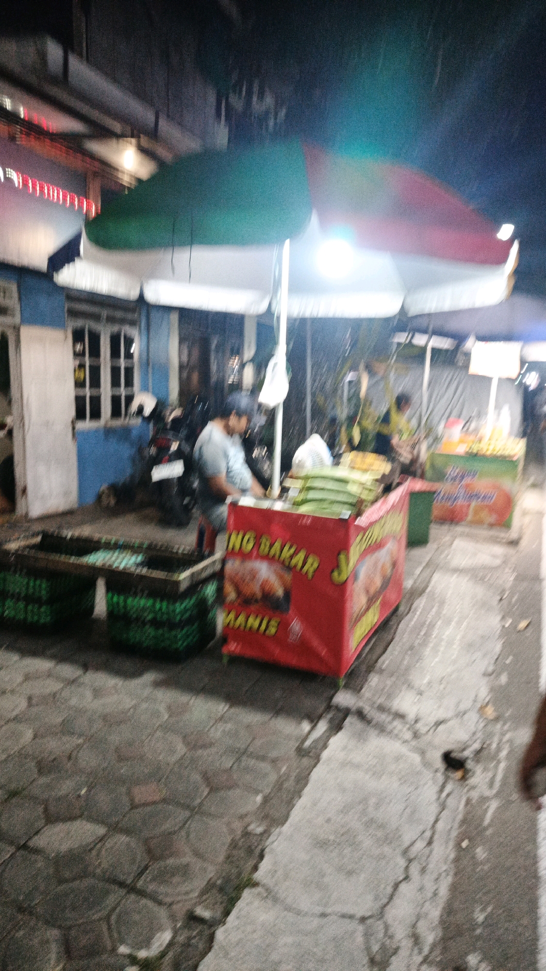 An outdoor scene at night in Surakarta, Indonesia, features a food stall. A person, likely the vendor, stands behind a red food cart adorned with text in Indonesian that reads "NO BAKAR MANIS." Green tarp-covered items are stacked on top of the cart. Adjacent to the cart are green plastic crates, one of which contains green food items. In the background, a blue building with a window is visible, along with a parked motorcycle. An umbrella, partially covering the stall, displays red, white, and green colors. The ground is paved with hexagonal stones. The scene is dimly lit, with some light sources creating glare.