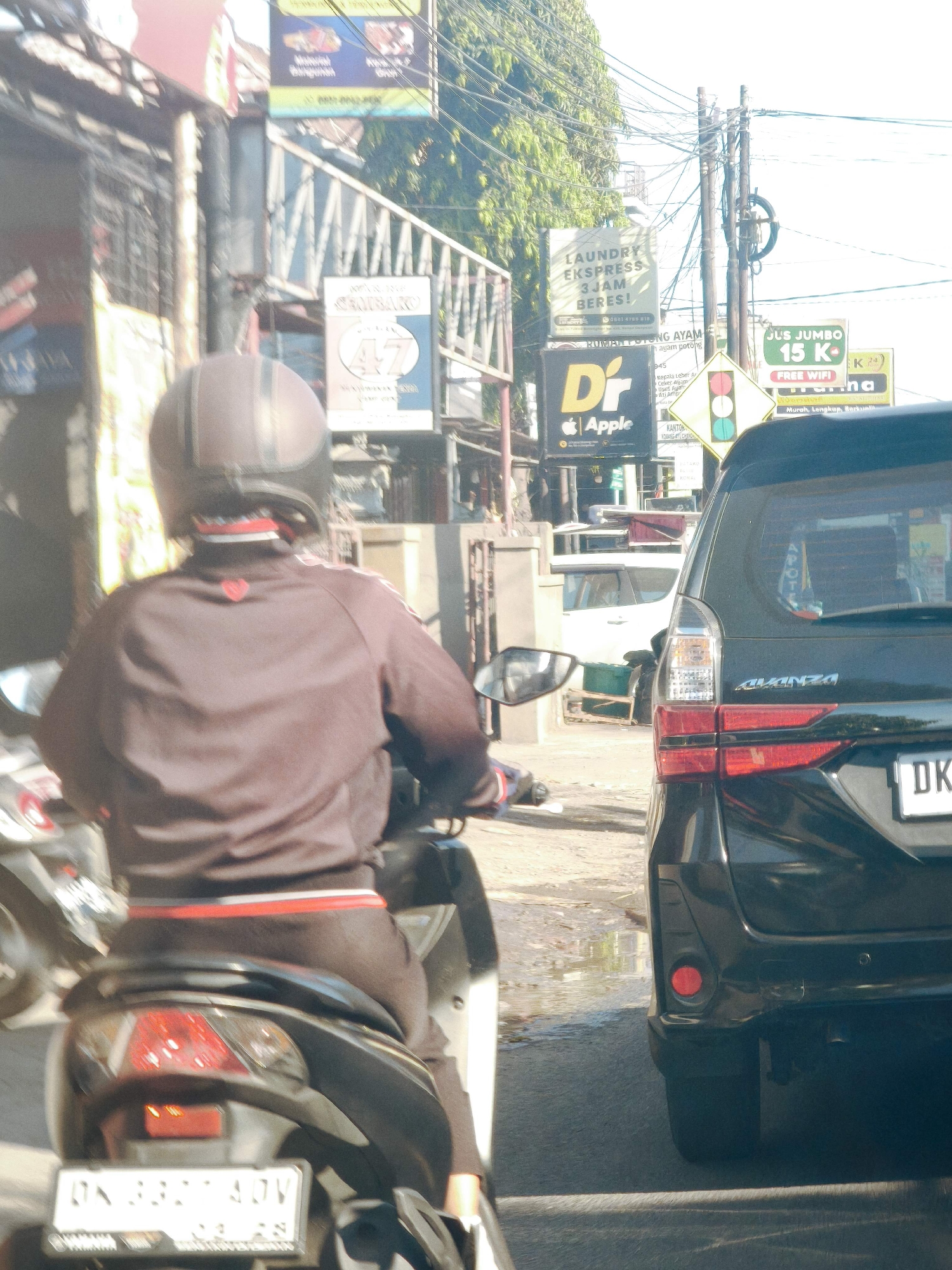 The image captures a street scene in Denpasar, Indonesia, likely on a busy road given the presence of multiple vehicles and advertisements. In the foreground, a person wearing a brown jacket and a dark helmet is riding a motorcycle. The motorcycle's license plate is partially visible and appears to read "DK 332 ADJV". To the right, a black Toyota Avanza is stopped, its rear visible. Several street signs are present, including one for "LAUNDRY EKSPRESS 3 JAM BERES!", another with "JUS JUMBO 15 K", and one displaying a traffic light symbol. The environment suggests a commercial area with shops and possibly residential buildings lining the street, indicated by the structures and overhead wires. The road surface appears wet in places, suggesting recent rain or cleaning. The overall lighting indicates it is daytime.