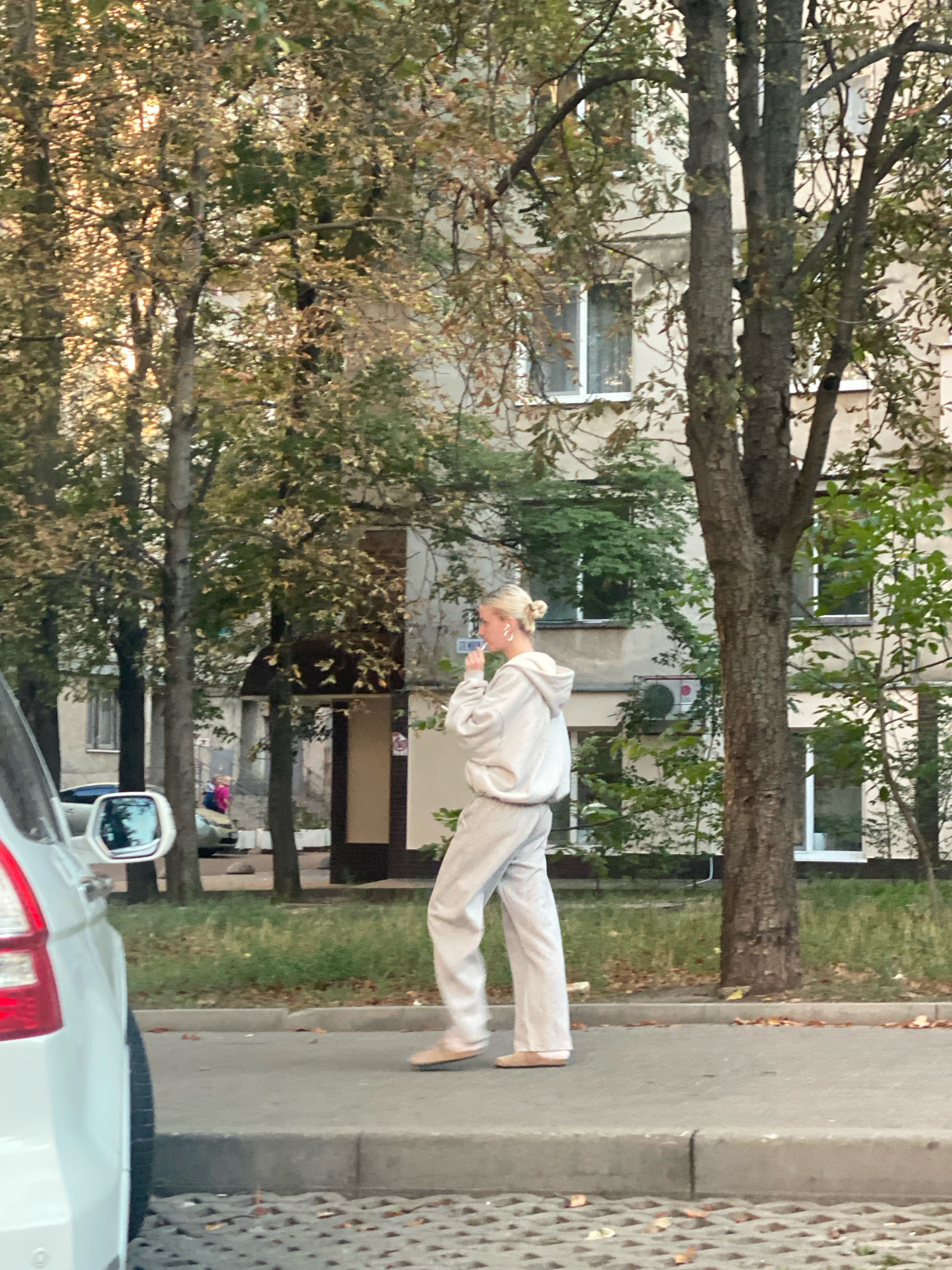An eye-level, medium shot shows a young woman with blonde hair pulled into a bun, wearing light-colored loungewear, walking on a sidewalk in Chișinău, Moldova. She is holding a phone to her ear, appearing to be engaged in a conversation. She is seen from the mid-torso down. The sidewalk is bordered by a curb and a grassy area with trees. In the background, a multi-story building with windows is visible, along with more trees. A white car is partially visible in the foreground on the left. The overall lighting suggests it is late afternoon or early evening, with sunlight filtering through the trees.