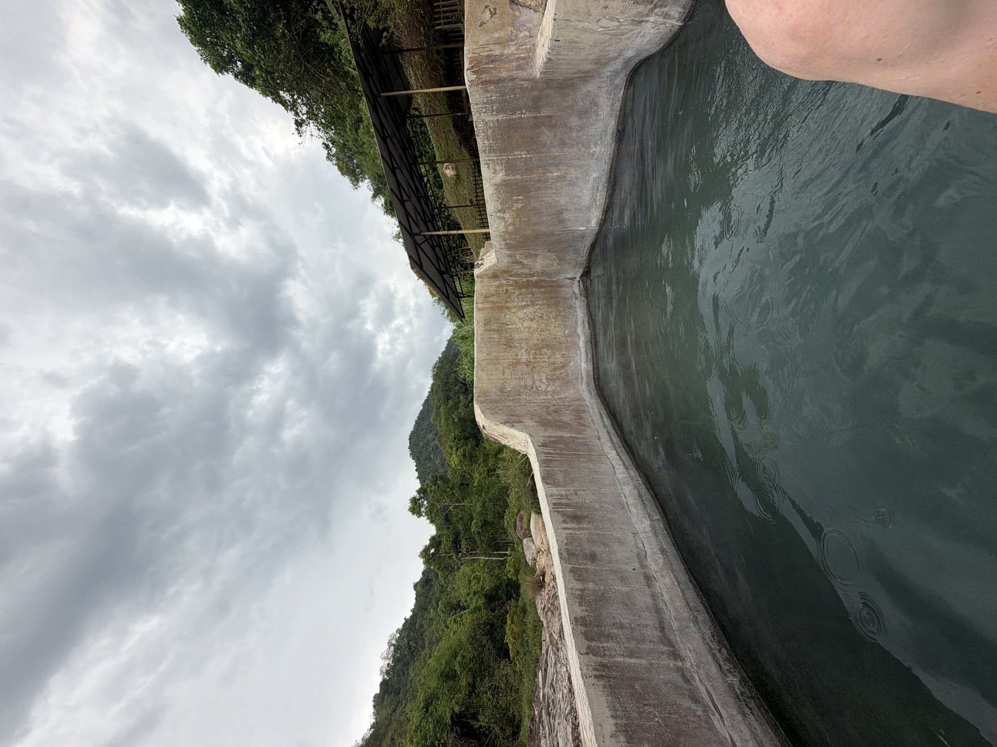 The image shows a man's knee and leg in the lower right corner, partially submerged in a dark, murky pool of water. The pool is contained within a concrete structure with rough, weathered walls. In the background, a lush, green hillside is visible, with a dense forest covering its slopes. A dark, open-sided shelter with a sloped roof stands on the hillside above the pool. The sky is overcast with heavy, grey clouds. The scene appears to be an outdoor hot spring or thermal pool located in the natural landscape of Khanh Vinh, Vietnam, given the tropical foliage and mountainous terrain.