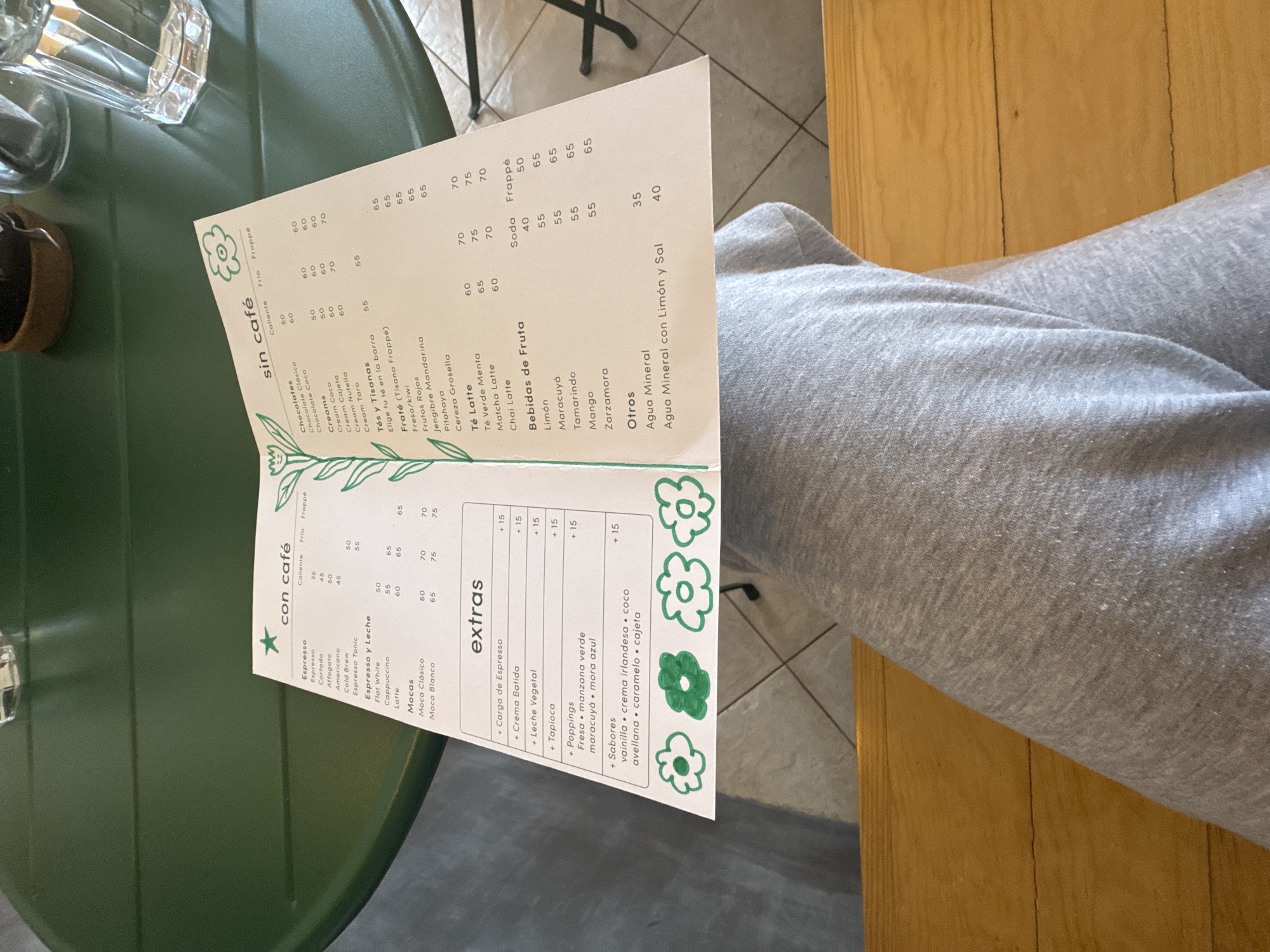 The image shows a menu with a green floral design, listing coffee, non-coffee drinks, and extras. The menu is in Spanish and includes items like "Espresso", "Latte", "Chocolate Clásico", and "Bebidas de Fruta". Prices are listed next to each item. The menu is resting on a green, possibly metal, table or surface. Part of a person's leg, clad in grey sweatpants, is visible in the upper right corner of the image. The menu is from a cafe in Oaxaca City, Mexico.