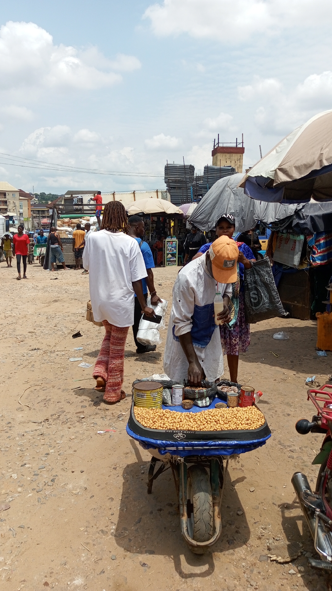 This media file captures a bustling outdoor market scene in Ogbunike, Nigeria, under a bright, cloudy sky. The ground is unpaved dirt, showing signs of foot traffic and minor debris like discarded plastic bottles.
In the foreground, a male vendor, wearing a white long-sleeved shirt with denim patches and an orange baseball cap, attends to his merchandise. He holds a white bottle (possibly containing a beverage) in his left hand while reaching into his goods with his right. His cart is a modified wheelbarrow, topped with a blue and black surface, displaying a large quantity of roasted peanuts, along with various canned items (red, yellow, and silver) and small bags. Text on the black border of his cart appears to read "KM SHAKM KANR SAN". Part of the text on his orange cap is visible, reading "STOP".
Behind the vendor, a woman in a patterned dress and a black baseball cap carries a black shopping bag with a white 'S' logo. To the left, a person with dreadlocks, dressed in a white oversized t-shirt and distinctive red and orange patterned trousers, walks away from the camera.
The background is filled with activity characteristic of a vibrant market. Numerous other people are visible, moving between makeshift stalls shaded by tarps and umbrellas of various colors (grey, black, beige, striped). A man in a red shirt is seen standing on top of a truck or similar structure in the mid-ground. In the distance, buildings and stacked construction materials (possibly scaffolding) are noticeable, indicating an urban or semi-urban setting. A red motorcycle is partially visible on the far right. The strong daylight suggests it is midday or early afternoon.