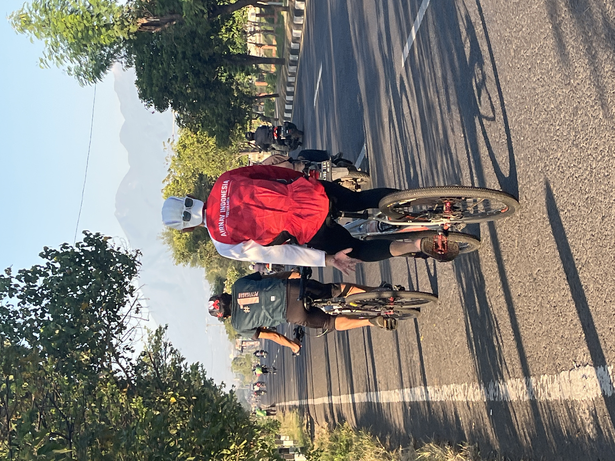 In the city of Siring, Indonesia, two individuals are engaged in a cycling activity along a paved road. The perspective is from behind the cyclists, capturing their movement. The cyclist in the foreground wears a red long-sleeved shirt with white accents and a white head covering, paired with white sunglasses. The shirt displays the text "AIRNAV INDONESIA YOGYAKARTA". To the left, another cyclist is seen wearing a dark green t-shirt with a white number "1" and the inscription "PETZESAGALAS" on the back, along with black cycling shorts. Both cyclists are on their bicycles, with the foreground cyclist on a bicycle with a silver frame and red detailing on the wheels. The background reveals a road lined with trees on both sides, and distant mountains under a clear blue sky. Several other cyclists and motorcycles are also visible on the road, indicating a group or event. The sunlight casts long shadows across the asphalt. The environment suggests a suburban or rural setting within the city, with a clear indication of a roadway designed for vehicle traffic.