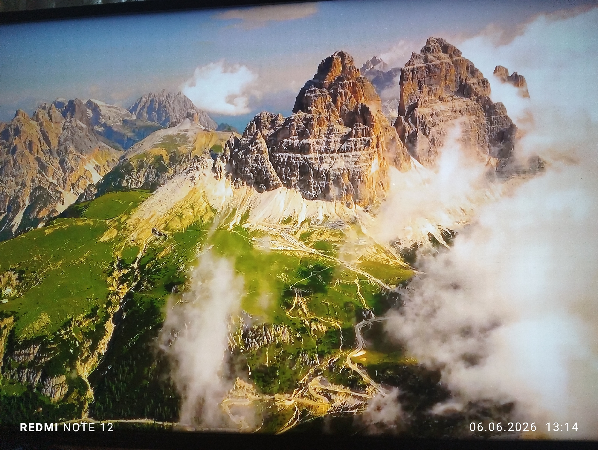 A photograph, captured by a Redmi Note 12 device in Burgas, Bulgaria, at 13:14 on June 6, 2026, depicts a rugged mountainous landscape displayed on a screen. The central focus is a series of towering, sharp, rocky peaks exhibiting shades of brown, orange, and grey, illuminated by sunlight. Lower slopes and valleys are covered in vibrant green vegetation, intersected by light-colored, possibly barren, areas and visible winding paths or roads. Wispy white clouds or mist partially obscure the lower portions of the mountains and valleys, particularly on the right side and in the foreground. The background features additional mountain ranges receding into a clear blue sky, which transitions to a lighter hue with some scattered clouds above the distant peaks. No people, animals, or artificial structures other than the aforementioned paths/roads are explicitly visible within the displayed landscape.