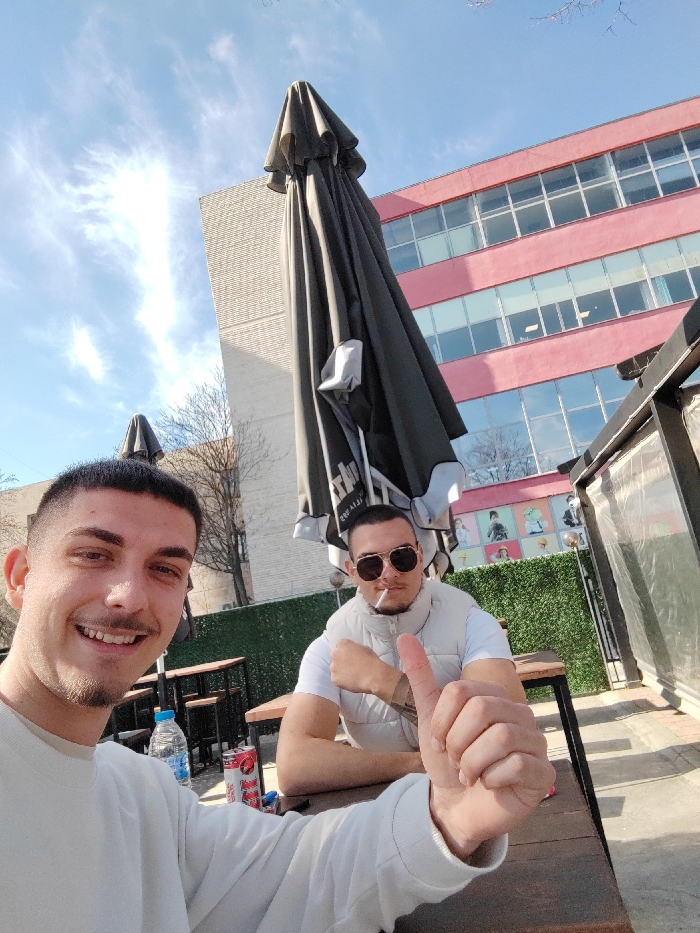 Two young adult males are depicted outdoors under a bright blue sky with wispy clouds, seated at a dark wooden table in what appears to be an open-air cafe or restaurant in Blagoevgrad, Bulgaria. The man in the foreground, positioned to the left, is smiling and looking directly at the camera, holding his right hand up in a thumbs-up gesture. He wears a white long-sleeved top and has short dark hair with a fade haircut and stubble. A clear plastic water bottle with a blue cap and a red and white can, identified as 'Monster,' are on the table in front of him. The man seated to the right wears dark round sunglasses, a white t-shirt, and a light-colored puffer vest, with a cigarette held in his mouth. His arms rest on the table, and he looks towards the camera. In the background, several large black closed patio umbrellas are visible. A prominent multi-story building with a distinctive reddish-pink facade and multiple horizontal rows of reflective windows, consistent with the American University in Bulgaria (AUBG) campus, is visible on the right. To its left, a lighter-colored building with vertical paneling stands. Bare tree branches are visible against the sky, and a green artificial hedge serves as a backdrop behind the second man.
