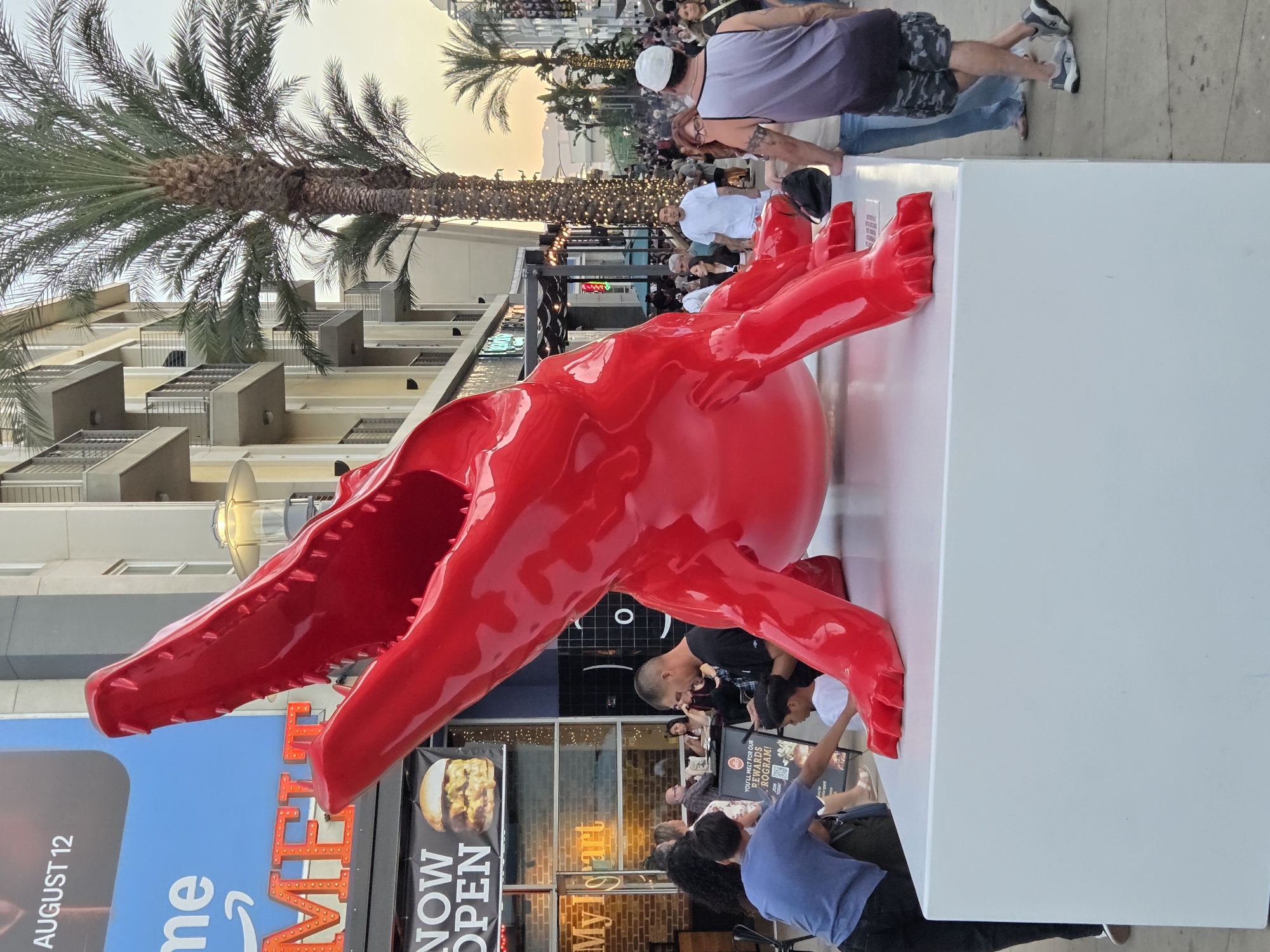 The image captures a scene in Burbank, United States of America, with a prominent, large, bright red sculpture of an alligator dominating the foreground. The alligator sculpture is positioned on a white, rectangular pedestal. Several people are visible around and near the sculpture. In the mid-ground, a man in a blue shirt and black pants is interacting with a sign, while another person, possibly a child, is reaching towards it. Behind them, several other individuals are gathered, some seated at tables outside what appears to be a restaurant or shop with the sign "NOW OPEN" and an image of a burger displayed. In the background, there is a multi-story building with balconies, palm trees adorned with lights, and a busy street with more people. The sky suggests it is late afternoon or early evening. The overall environment appears to be an outdoor public space, possibly a commercial or entertainment district.