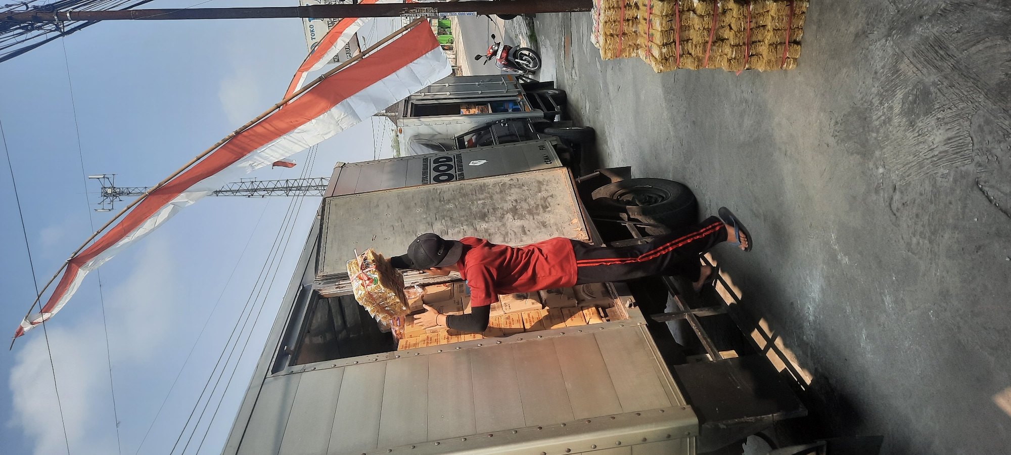 An Indonesian man wearing a red shirt, black pants with red stripes, and a black cap is loading stacked boxes into the back of a silver truck in Selopuro, Indonesia. He is reaching up with a stack of yellow packaged goods. The truck is parked on a concrete surface, and there are more stacks of the same yellow packaged goods to the right of the truck. The scene is outdoors during the day, with a clear sky and some power lines overhead. A red and white Indonesian flag is flying diagonally across the upper portion of the image. Another truck is parked behind the first one. A red motorcycle is parked to the right of the second truck.