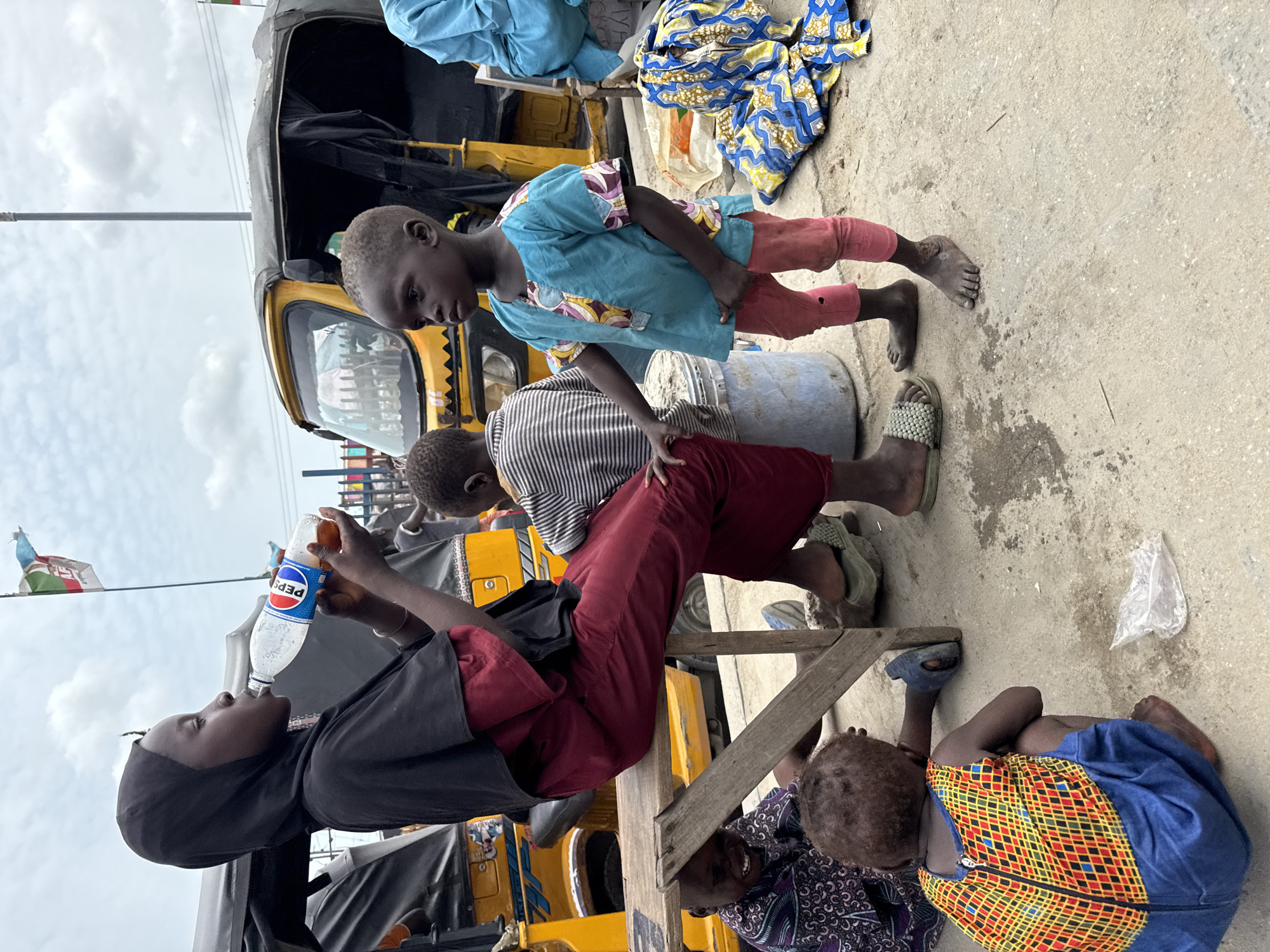 This image captures an outdoor scene in Lekki, Nigeria, featuring several young individuals and children on what appears to be a concrete street or market area, under an overcast sky.
In the foreground, a young person, likely a girl, wearing a black hijab and dark red pants, is seated on a low wooden bench and is drinking from a clear plastic Pepsi bottle. Her gaze is directed upwards, focusing on the bottle. To her right, a child in a light blue top with patterned sleeves and pink pants stands barefoot. Next to the standing child, another child in a striped top and dark red pants is also seated, leaning against an unseen object. Further to the right and lower, a young child wearing a vibrant blue dress with a yellow, red, and black patterned front is sitting or lying on the concrete ground. Another child, partially visible, wearing a purple patterned top, is also present near the wooden structure.
In the background, a yellow auto-rickshaw, commonly known as a keke napep or tuk-tuk, is parked. Bundles of colorful fabric are visible near the vehicle. The ground is a light-colored concrete, showing signs of wear. The visible text is "PEPSI" on the bottle. The scene depicts a casual moment of daily life, with children present in a public, potentially bustling environment.