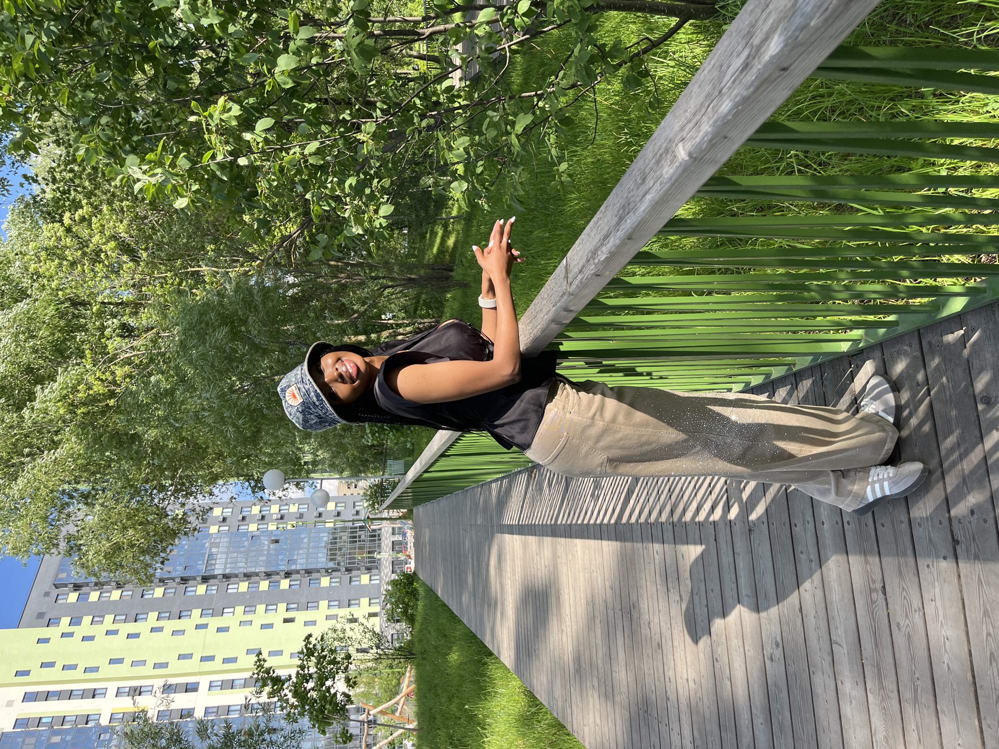 A young Black woman is standing on a wooden boardwalk, leaning against a green metal railing. She is wearing a black sleeveless top, tan wide-leg pants adorned with rhinestones, a patterned bucket hat, and white sneakers. The woman is smiling and looking to her right. Behind her, there is lush green grass, trees, and a tall, modern building with light green and grey facades. The boardwalk extends into the distance. This scene appears to be in a park or recreational area within the city of Kazan, Russia, given the architectural style of the building and the surrounding urban landscaping.