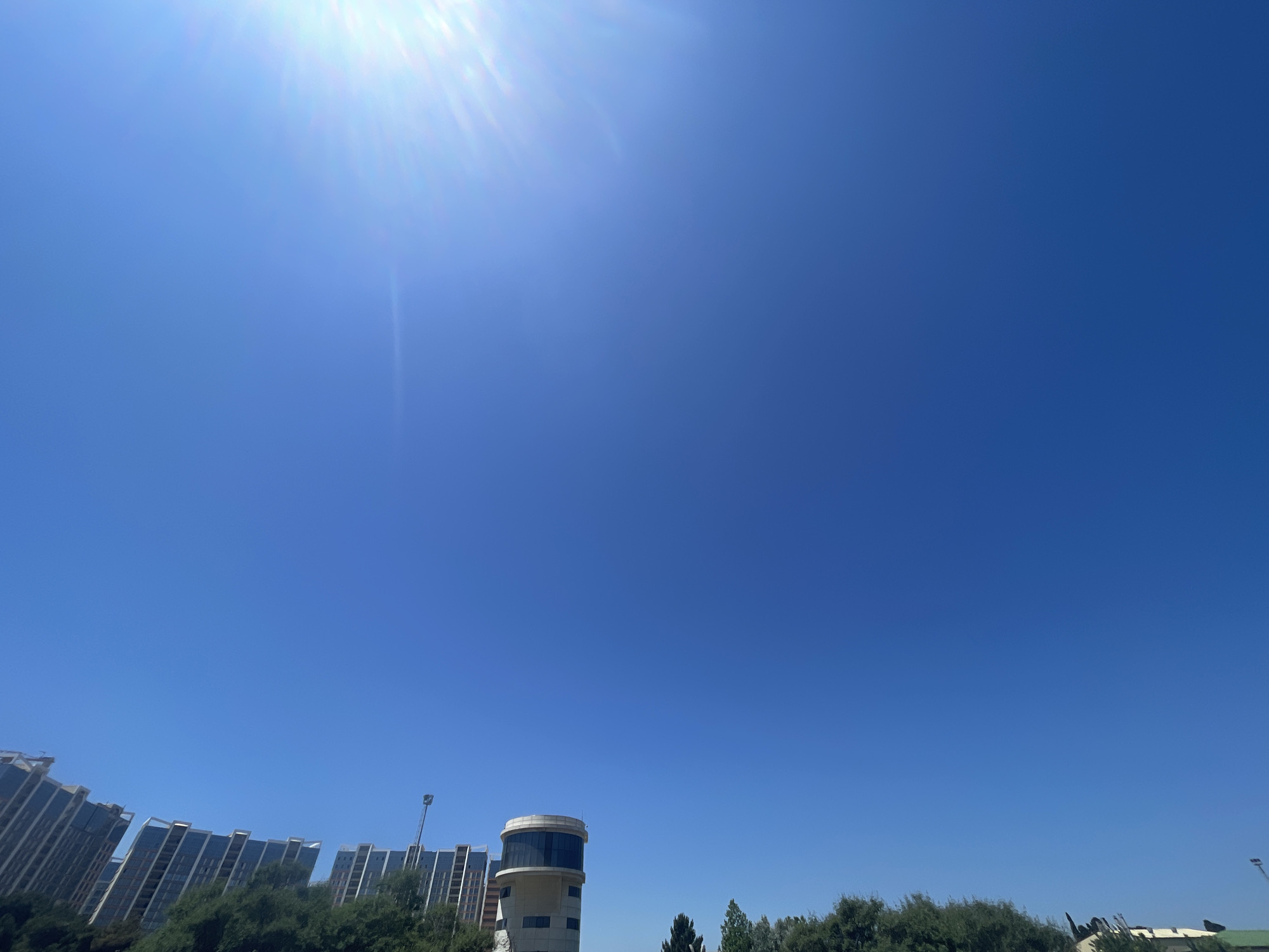 The media file captures a clear, bright sky with the sun prominently featured in the upper left corner, casting lens flare. The scene is set in Baku City, Azerbaijan, and depicts a cityscape below the expansive blue sky.
In the lower portion of the image, a row of modern apartment buildings with distinctive architectural features is visible, characterized by their reflective facades and angled designs. To the right of these buildings, a unique, cylindrical tower structure with large windows stands out. This tower might be a water tower or a building with a specific function, like an observation deck. Dense green foliage of trees lines the foreground, partially obscuring the lower parts of the buildings.
The bright sunlight and clear sky indicate it is daytime, likely mid-morning or early afternoon, under fair weather conditions. There are no people visible in the frame, and the focus is on the architecture and the natural elements. The overall impression is one of a modern urban environment bathed in the warm light of a sunny day. No visible text is present in the image.