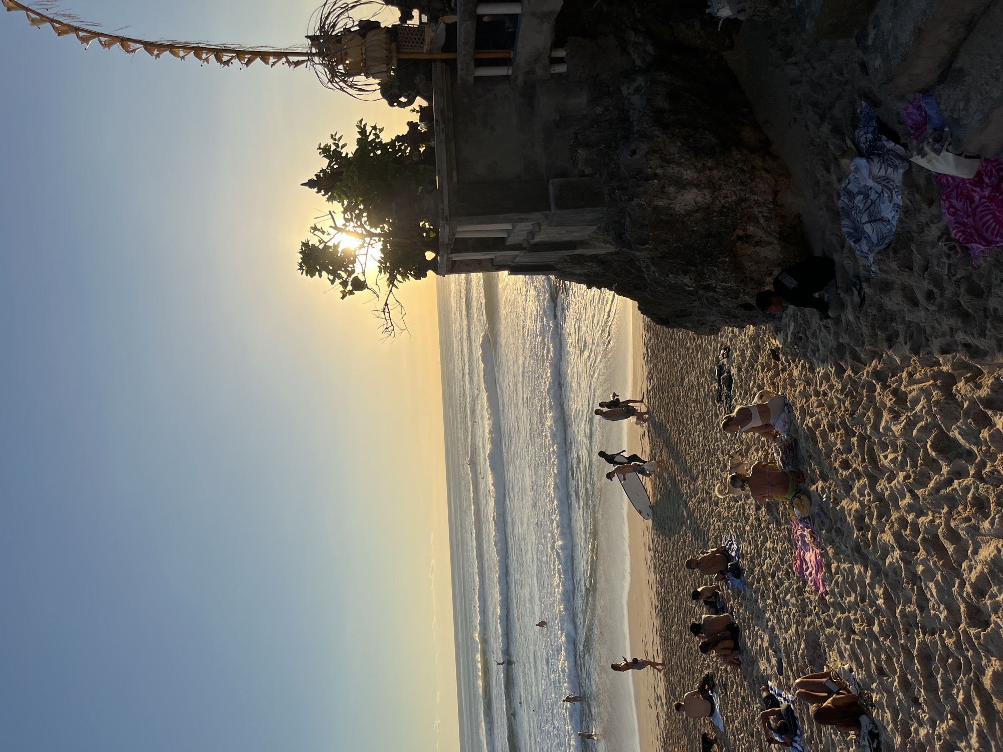 At sunset on a beach in Kuta Selatan, Indonesia, a crowd of people relax on the sand while the sun casts a warm glow across the ocean. The waves are gentle, and a few surfers can be seen in the distance. On the right side of the frame, a stone structure sits on a cliff overlooking the water, adorned with traditional Balinese decorations. The beach is sandy, with people scattered across it, some lying down, others sitting and chatting. Several beach towels and blankets are spread out on the sand, and a few dogs are present. The sky is clear, with a soft gradient of blue and yellow as the sun begins to dip below the horizon.