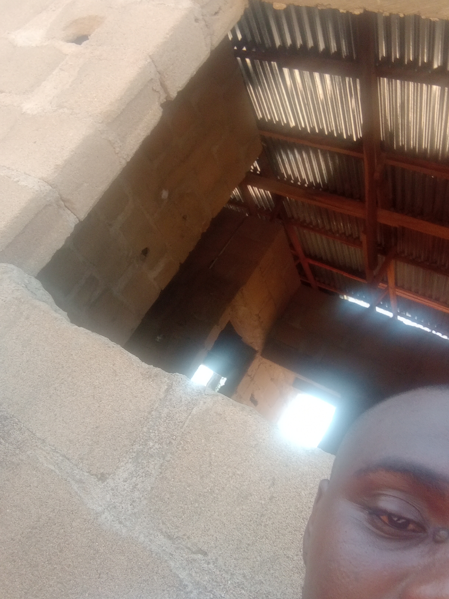 The image shows a close-up of a person's face, partially obscuring the view of an unfinished or dilapidated structure. The structure appears to be made of concrete blocks, with a corrugated metal roof supported by wooden beams. Light streams in from an opening in the structure. The person's face is dark-skinned, and they have dark eyes and a shaved head. The scene appears to be outdoors, with the harsh sunlight suggesting a warm climate. The overall impression is one of poverty or neglect.