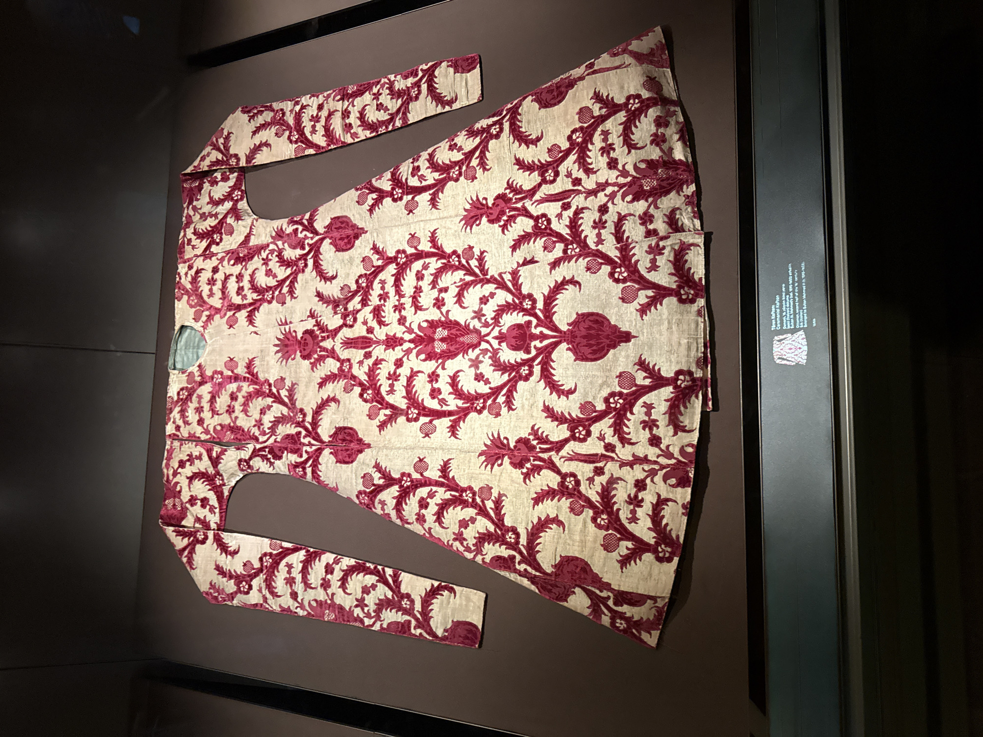 A long-sleeved, embroidered kaftan with a repeating pattern of red floral motifs on a cream-colored background is displayed in a museum exhibit. The kaftan has a V-neckline and flares out towards the bottom. A label beneath the kaftan reads "Teren Kaftan", followed by information in Turkish and English describing the garment as belonging to Sultan Mehmed IV. The exhibit appears to be located in Istanbul, Turkey.