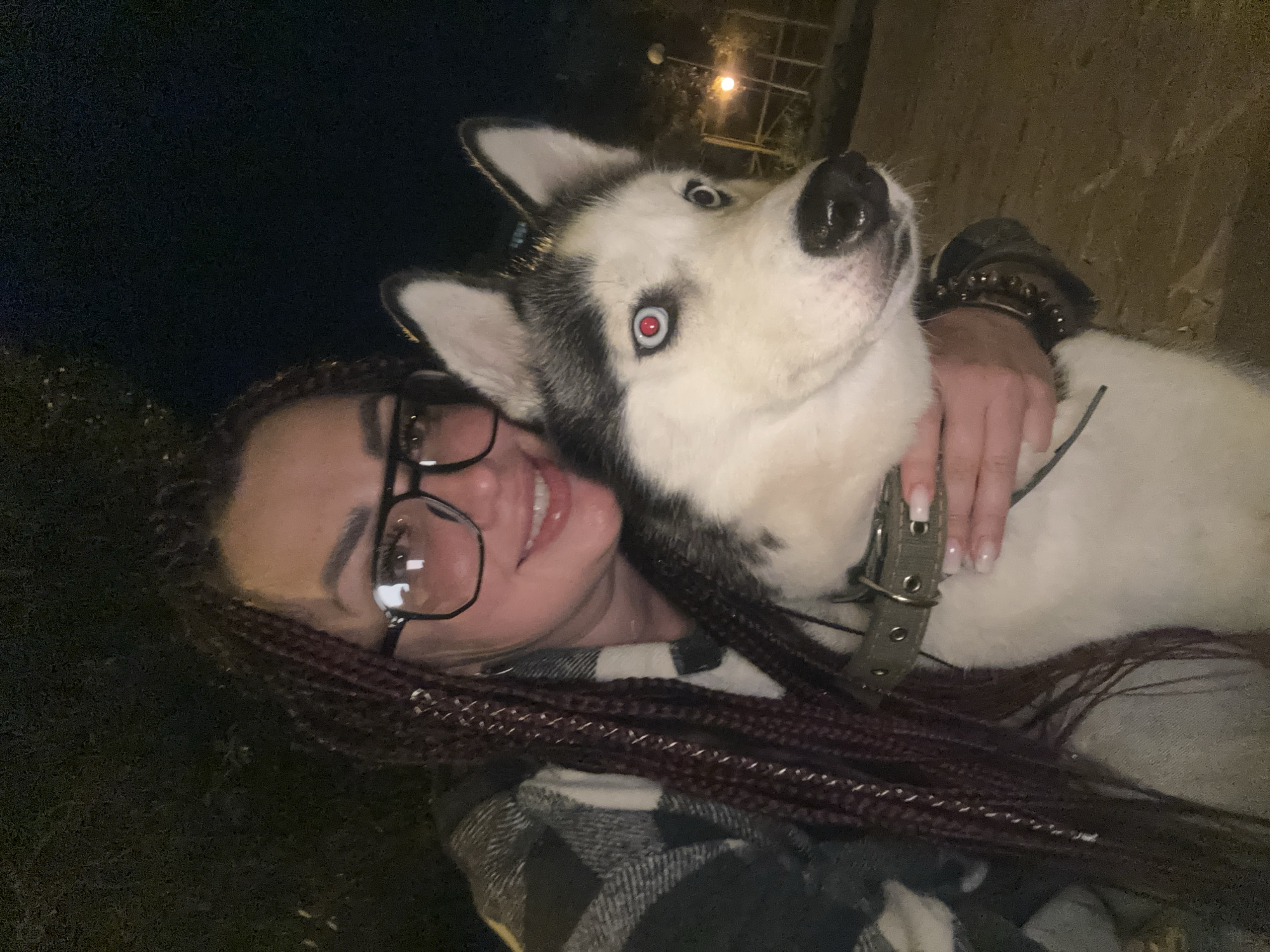 The media file is a selfie of a young woman with auburn braids and glasses posing with a husky dog at night. The woman is smiling, and the dog has its head close to her face, with its blue eyes looking directly at the camera. The background is dark, with some faint lights visible, suggesting an outdoor setting at night. The woman is wearing a plaid shirt. The dog has a green collar with metal studs. The overall mood is affectionate and playful, with both subjects appearing happy. There is no visible text in the image. The location context is Tolyatti, Russia.