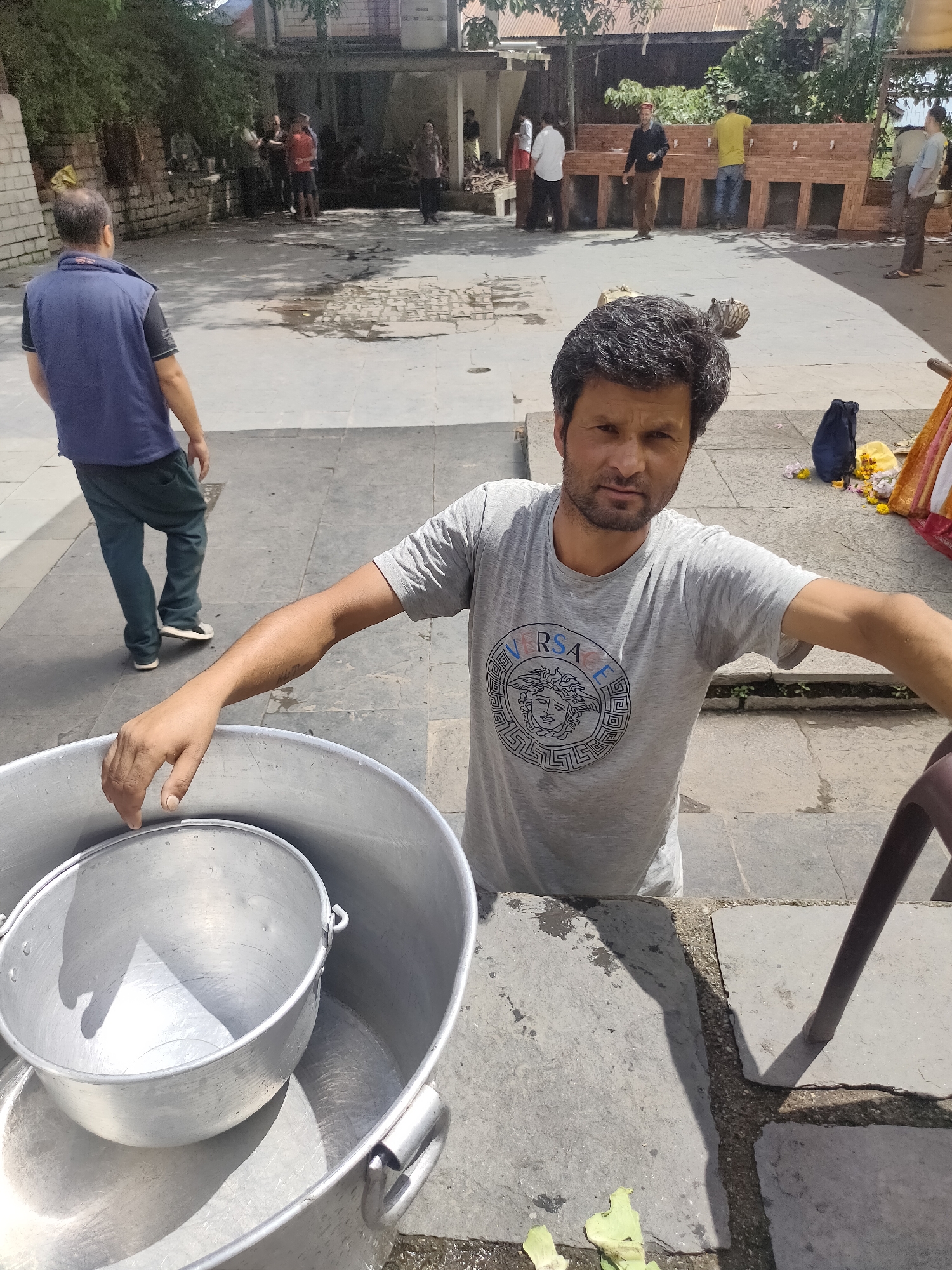 In Siyal, India, a man stands outdoors, looking towards the camera. He is wearing a grey t-shirt with a graphic print of the Versace logo. His right arm is extended, resting on the rim of a large metal pot. Inside this pot is a smaller metal pot. Behind the man, there is a paved courtyard with several people in the background, some near a row of sinks and others walking. To the left, a man in a blue vest and dark pants is walking away from the camera. The environment appears to be a public space, possibly a temple complex or a communal area, with stone paving and some greenery.