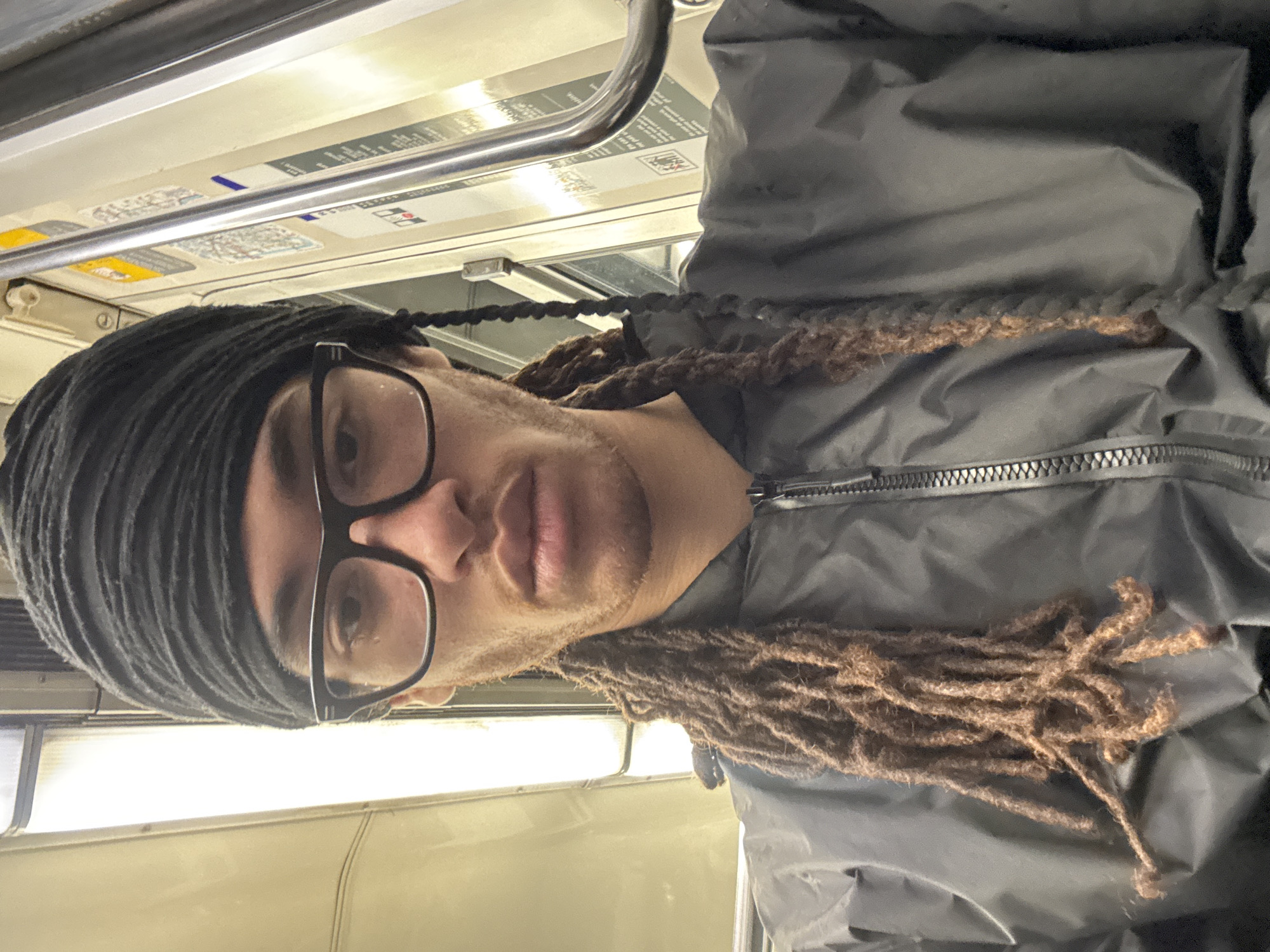 A selfie taken indoors, likely in a Paris Metro subway car. A person with brown dreadlocks partially covered by a black headwrap, wearing black-rimmed glasses and a dark grey jacket, is looking at the camera. The interior of the subway car is visible in the background, featuring a handrail, overhead information panels, and a window.