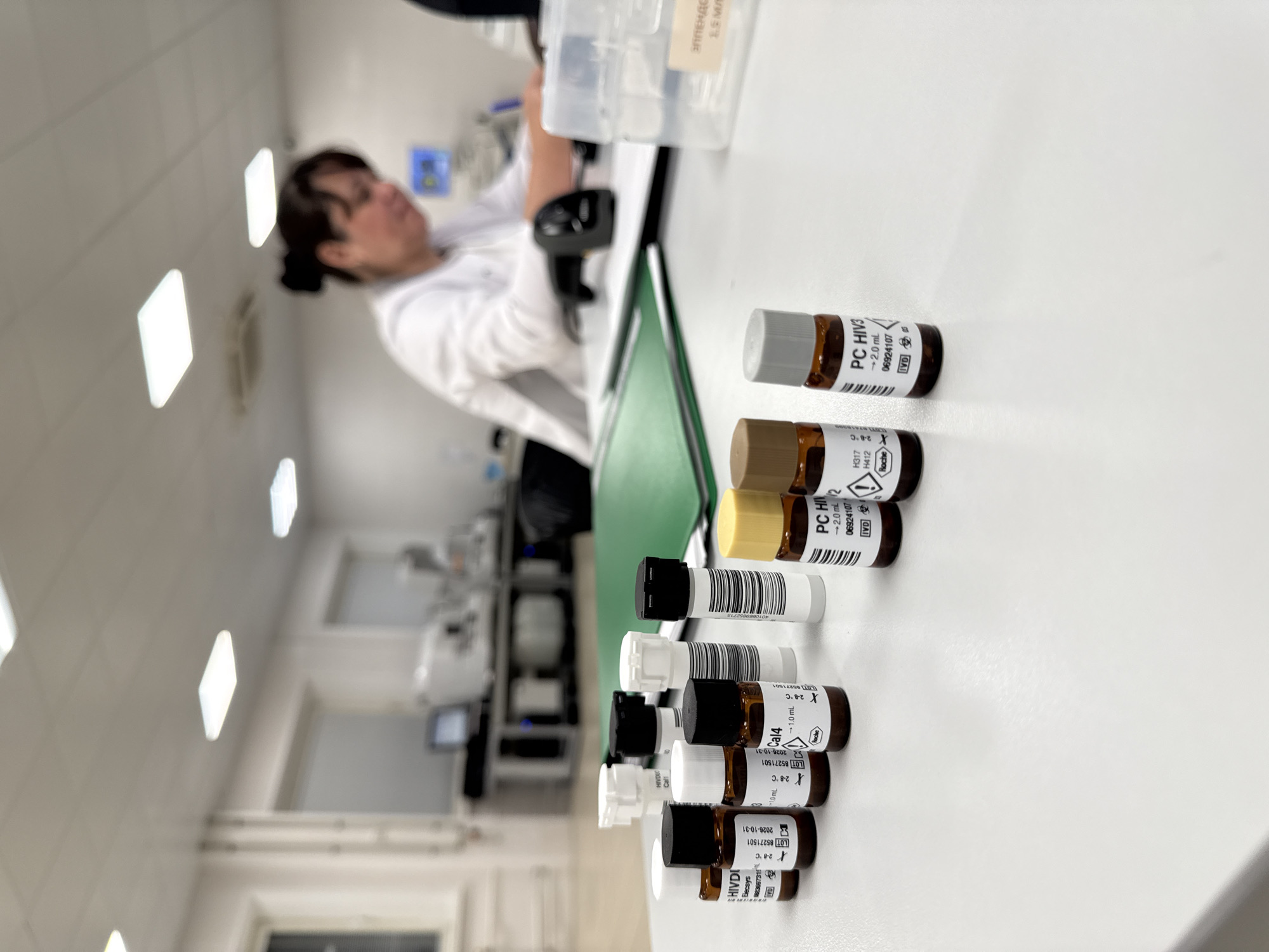 The image depicts a laboratory setting, possibly a medical research or testing facility, with a clear foreground and a blurred background. In the foreground, there are several small glass vials with stoppers, arranged on a white surface. Some of these vials have labels with text and barcodes, indicating they contain reagents. One vial is labeled "HIVDI" and "Cal4." Another vial is labeled "PC HIV2" and a third with "PC HIV3." Behind the vials, a green folder lies open. Further in the background, a person wearing a white lab coat is seated at a desk, appearing to be writing or working with documents. Various pieces of laboratory equipment, including what look like centrifuges and monitors, are visible in the blurred background. A barcode scanner is present on the desk in front of the person. The environment appears to be indoors, with overhead lighting. There are no overt indicators of the specific location within Moscow, Russia, nor are there any details suggesting the time of day or weather conditions. The scene suggests an ongoing scientific or analytical process.