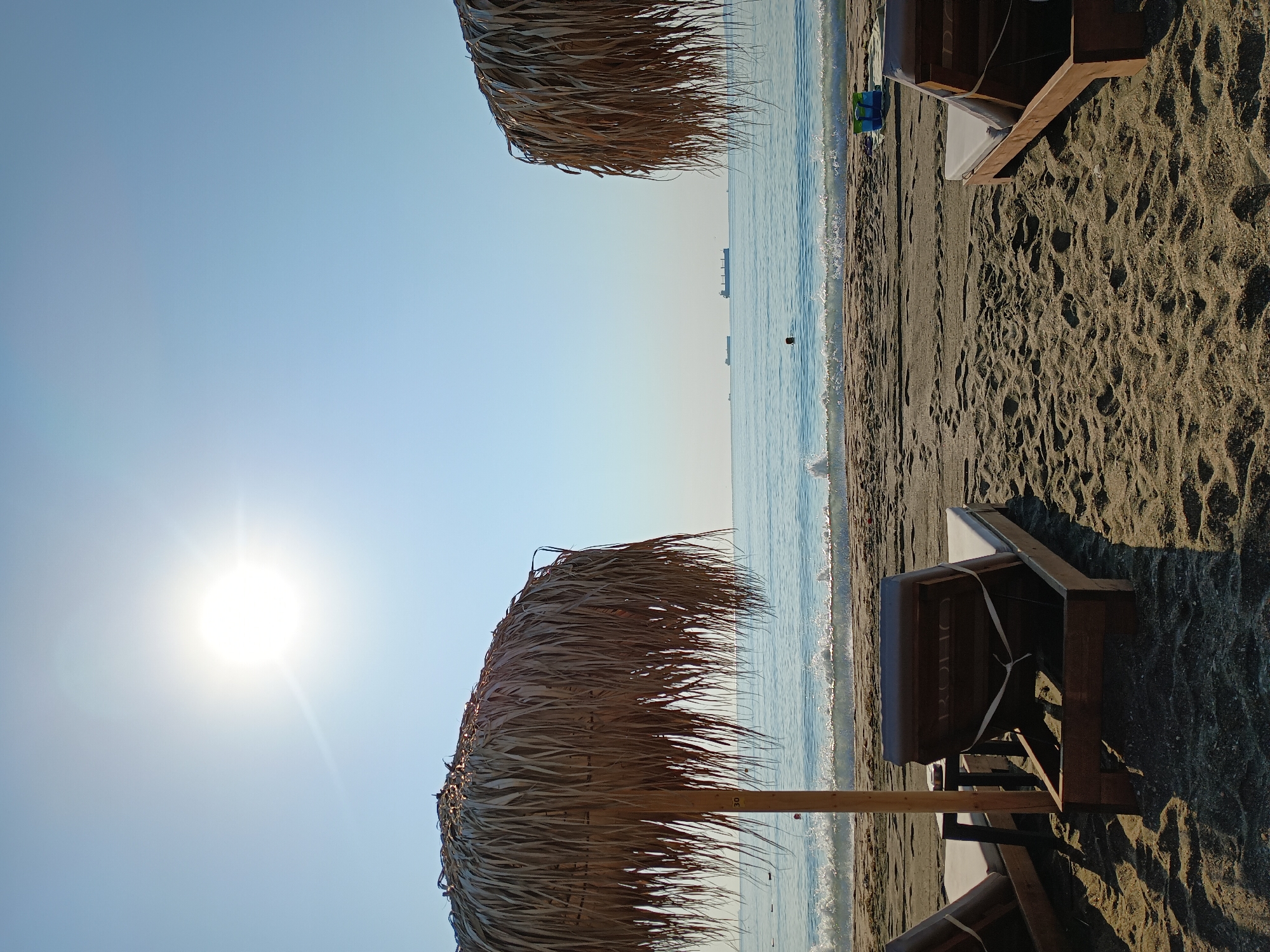 The image depicts a serene beach scene, likely on the Black Sea coast near Burgas, Bulgaria, judging by the presence of sun loungers and beach umbrellas. The sun is high in the sky, casting a bright glare and creating long shadows on the sandy beach. Two thatched beach umbrellas dominate the foreground, with one on the left and another partially visible on the right. Several wooden sun loungers are arranged on the sand, some occupied by cushions. The gentle waves of the sea are breaking on the shore, and in the distance, several cargo ships can be seen on the horizon. A lone swimmer is visible in the water. The overall atmosphere is one of peaceful relaxation and a perfect summer day.