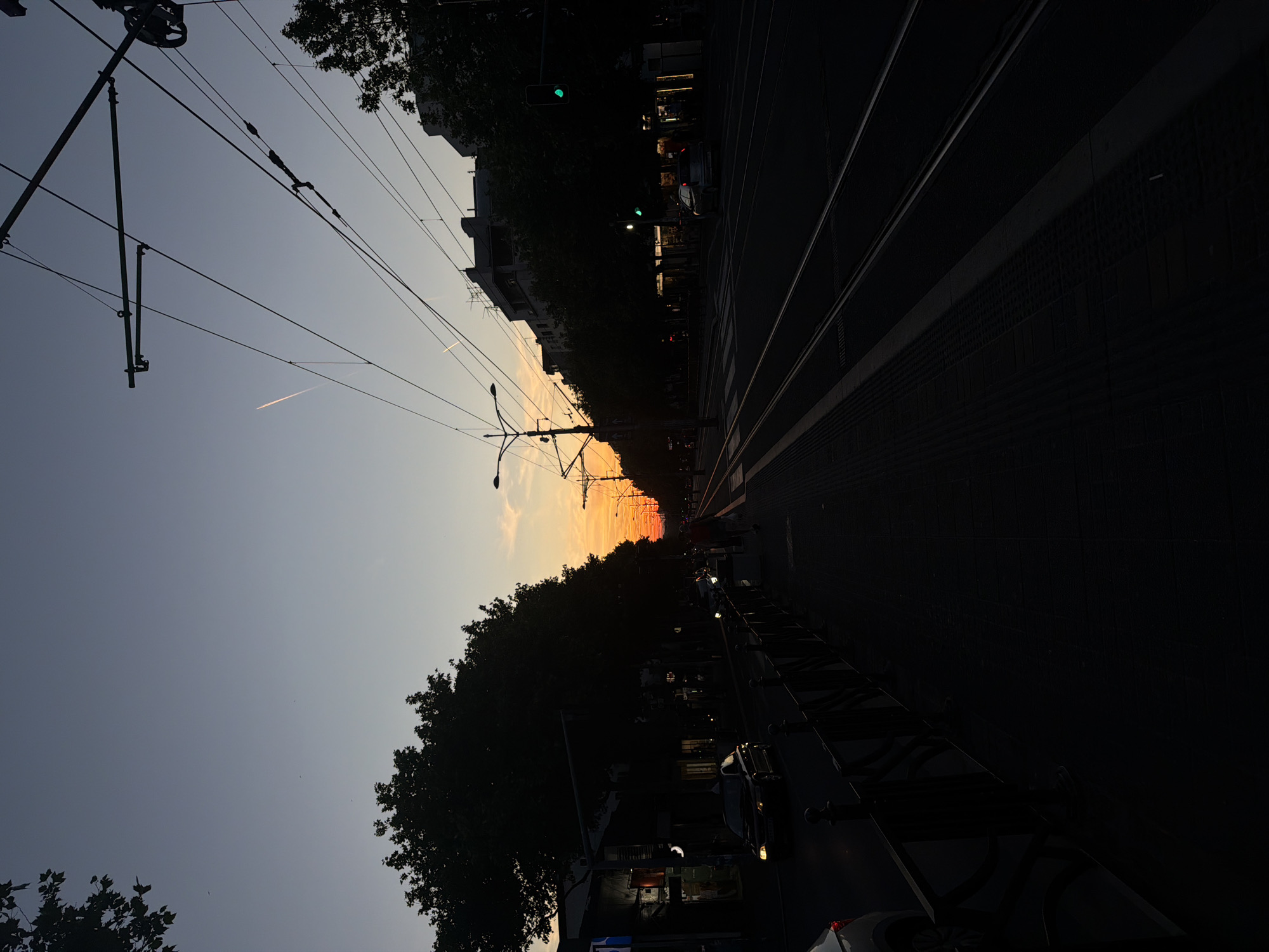 This image captures a vibrant urban street scene at sunset, viewed from a perspective slightly below the overhead wires and looking down the street. The sky dominates the upper half, transitioning from a light blue-grey on the left to striking orange and yellow hues at the horizon, where the sun is setting. A faint white contrail trails across the upper-left sky.
A dense network of black overhead power lines and tram wires crisscrosses the sky, forming intricate patterns and appearing as stark silhouettes against the lighter backdrop. Along the left side of the street, a continuous row of trees stands as a dark silhouette, obscuring the lower parts of multi-story buildings that line the street.
The right side of the image features the street itself, with prominent tram tracks running parallel into the distance and converging towards the colorful horizon. The road surface is dark, suggesting the dimming light of dusk. Scattered along the street are a few vehicles, primarily appearing as dark, shadowy forms. A green traffic light is visible on the left side of the road, further down, and another appears in the distance. The overall atmosphere is one of peaceful transition from day to night in a bustling city environment. No discernible people or readable text are visible.