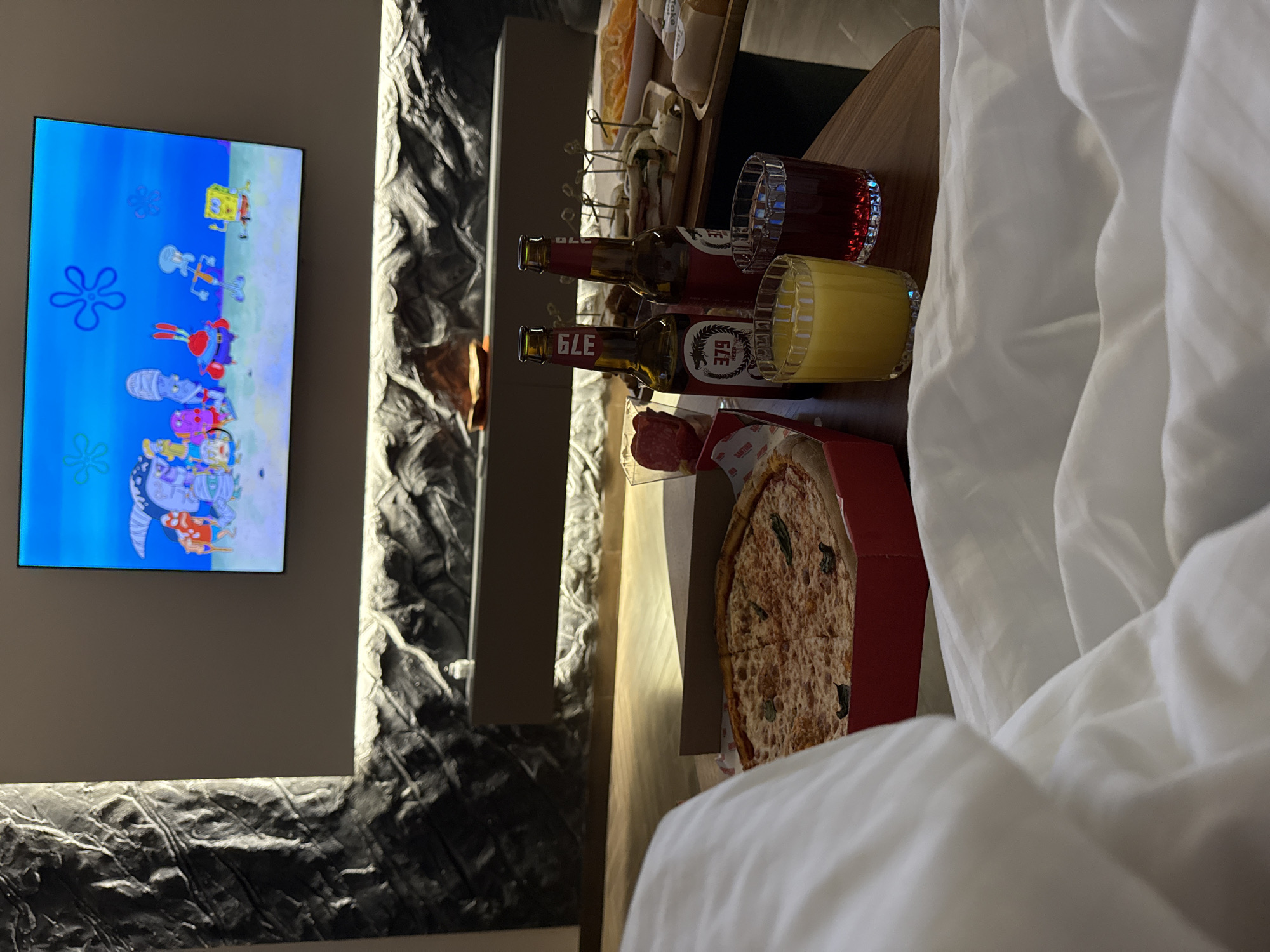 An indoor scene depicts a comfortable leisure setting, likely a hotel room in Yerevan, Armenia. The primary activity involves relaxing and enjoying food and beverages while watching television.
In the foreground, white bedding, possibly a duvet or sheets, is visible, suggesting someone is in bed or relaxing nearby. To the right, a wooden surface serves as a tray or table, holding an assortment of food and drinks. There are two beer bottles, both featuring the number "379" and one clearly labeled "GLE Beer." Two glasses are present: one filled with a red liquid and another with a yellow liquid, likely fruit juice. A large, round pizza, topped with green herbs, is in a red delivery box, with a slice already removed. Faintly visible text on the pizza box lining appears to be "SANTINO." Further back on the table, another platter of snacks or appetizers, possibly small sandwiches or canapés on skewers, can be seen.
On the left, a flat-screen television is mounted on a wall, displaying a scene from the animated cartoon "SpongeBob SquarePants." Visible characters include SpongeBob, Mr. Krabs, and others in a colorful underwater setting. The wall around the TV features a textured, possibly stone or sculpted, dark gray material, illuminated by indirect lighting, suggesting an evening or nighttime setting. The overall atmosphere is one of relaxation and indulgence.