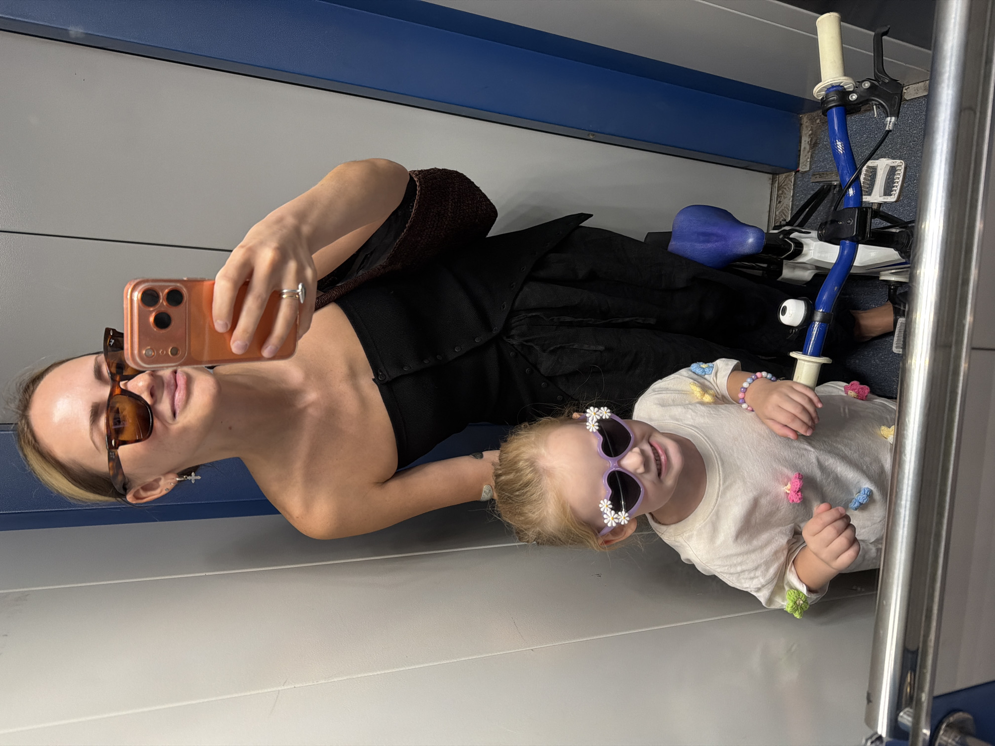 A woman and a young child are captured in a mirror selfie within a confined space, resembling an elevator or a public transport cabin.
The woman, positioned behind and to the right of the frame, has blonde hair styled with some strands framing her face. She wears brown tortoise-shell framed sunglasses with amber-tinted lenses. She is dressed in a black strapless top and black loose-fitting trousers with visible drawstrings. Her left ear features a silver hoop earring with a small cross charm. On the ring finger of her left hand, she wears a silver-colored ring. She holds an orange-cased smartphone with a triple-lens camera module in her left hand, taking the photograph. A dark brown, textured (possibly knitted or crocheted) shoulder bag is draped over her left shoulder. She is smiling slightly.
The child, standing in front and slightly to the left in the foreground, has blonde hair tied back. She wears lavender heart-shaped sunglasses adorned with white daisy appliques on the upper corners. She is smiling broadly, showing her teeth. She wears a light-colored (off-white or cream) long-sleeved t-shirt decorated with multiple colorful flower appliqués in shades of pink, green, yellow, and blue. Several colorful beaded bracelets are visible on her right wrist.
A child's bicycle with a white frame, blue handlebars, and a blue seat is positioned to the right of the child. The handlebars feature white grips and a small white bell.
The background consists of light gray wall panels on the left and a darker gray panel on the right, which features a distinct vertical blue strip, suggesting a door frame or structural element. The floor at the bottom of the frame appears to be a textured, possibly ribbed, surface. There is no discernible information within the image to identify the specific location as Khimki, Russia.